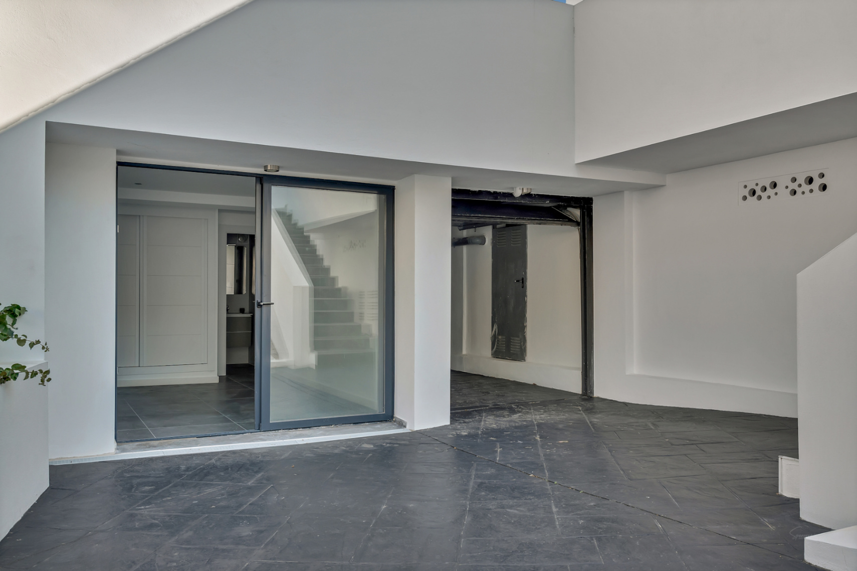 Ingang op de benedenverdieping met glazen schuifdeur, donkere tegelvloer en een functionele garage- of bergruimte in een strakke witte architectuur.