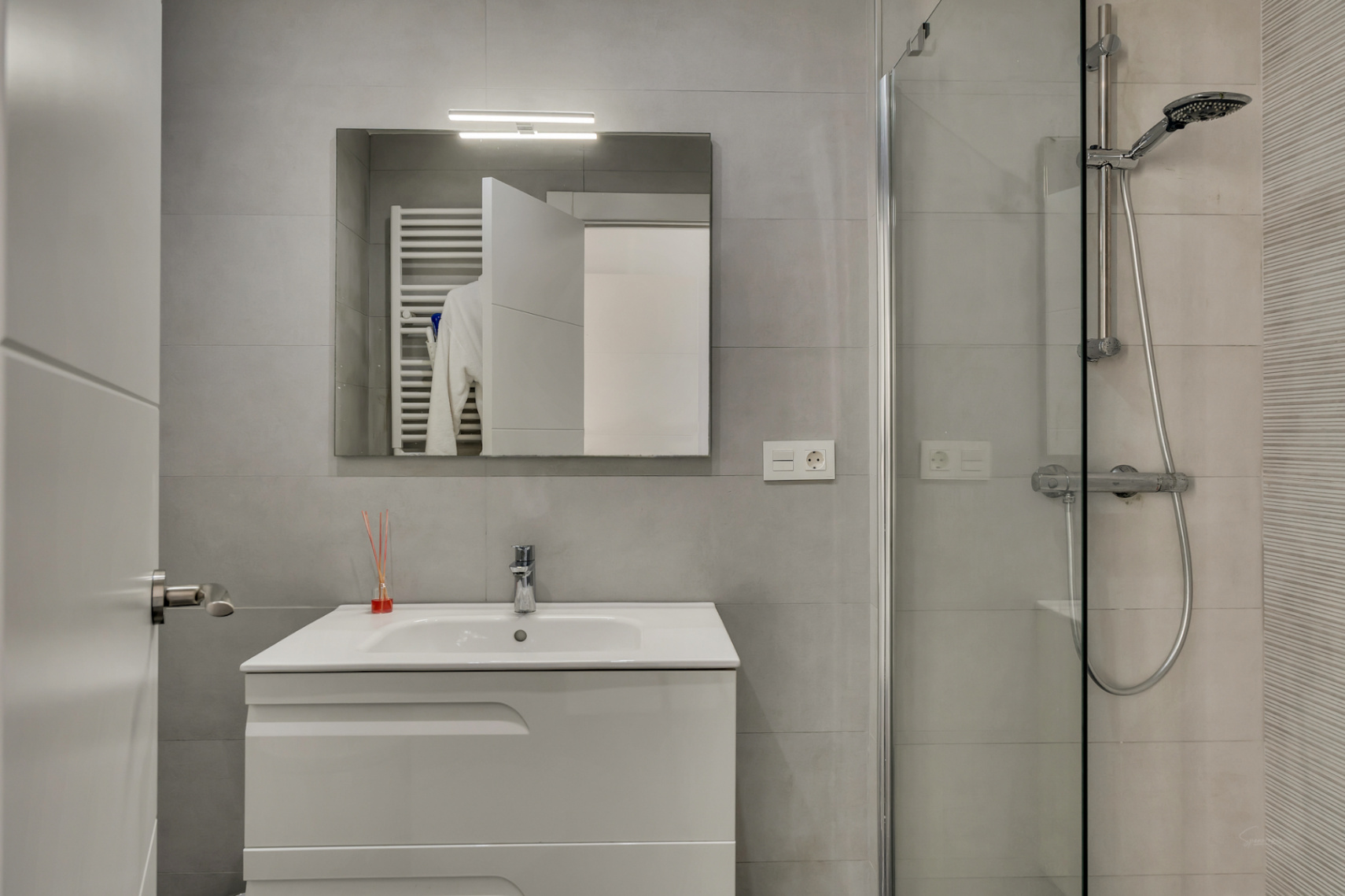 Moderne badkamer met grijze tegels, een wit wastafelmeubel, een verlichte spiegel en een inloopdouche met glazen wand en chromen kranen.
