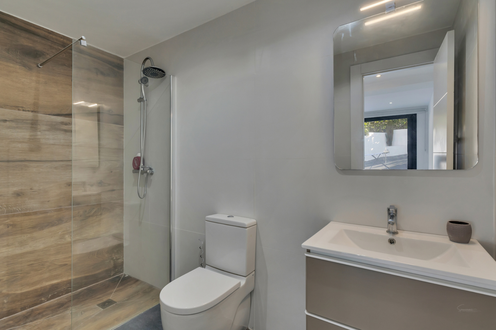 Moderne badkamer met inloopdouche voorzien van houtlook tegels, glazen wand, wit toilet en wastafelmeubel met een spiegel met geïntegreerde verlichting.