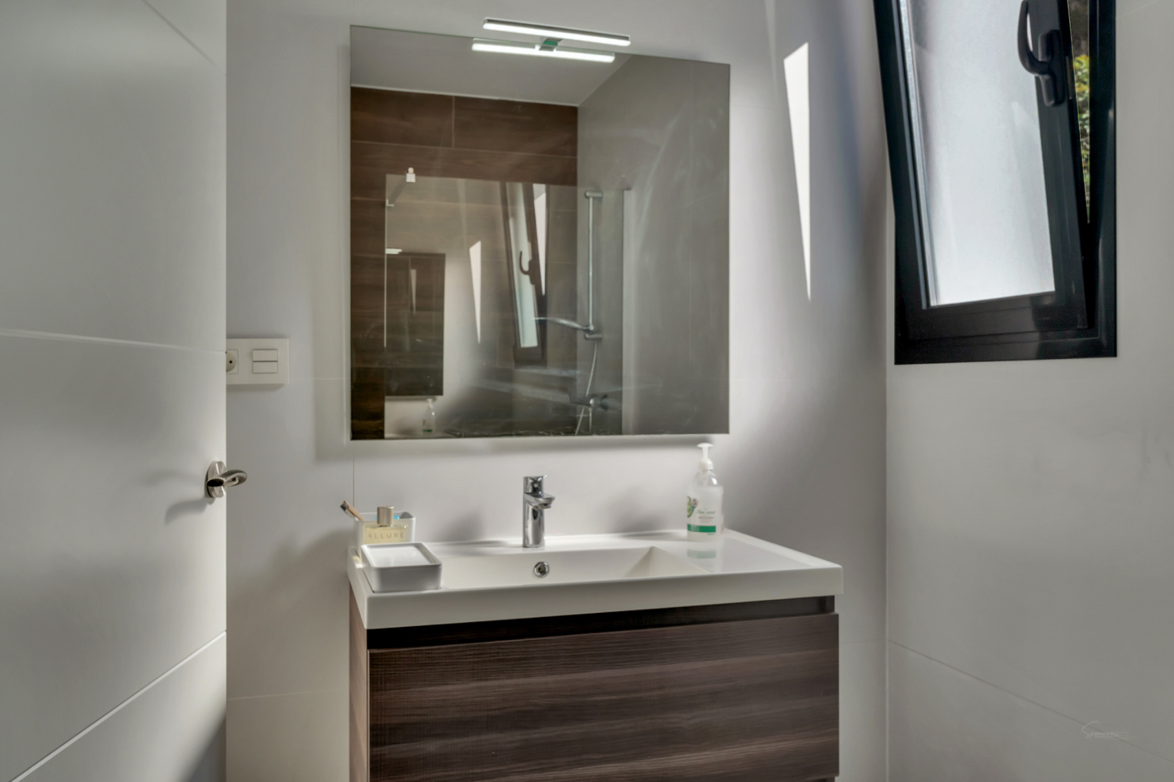 Moderne badkamer met zwevend wastafelmeubel in donker hout, witte wasbak en spiegel met bovenverlichting. Voorzien van een zwart geëmailleerd raam.