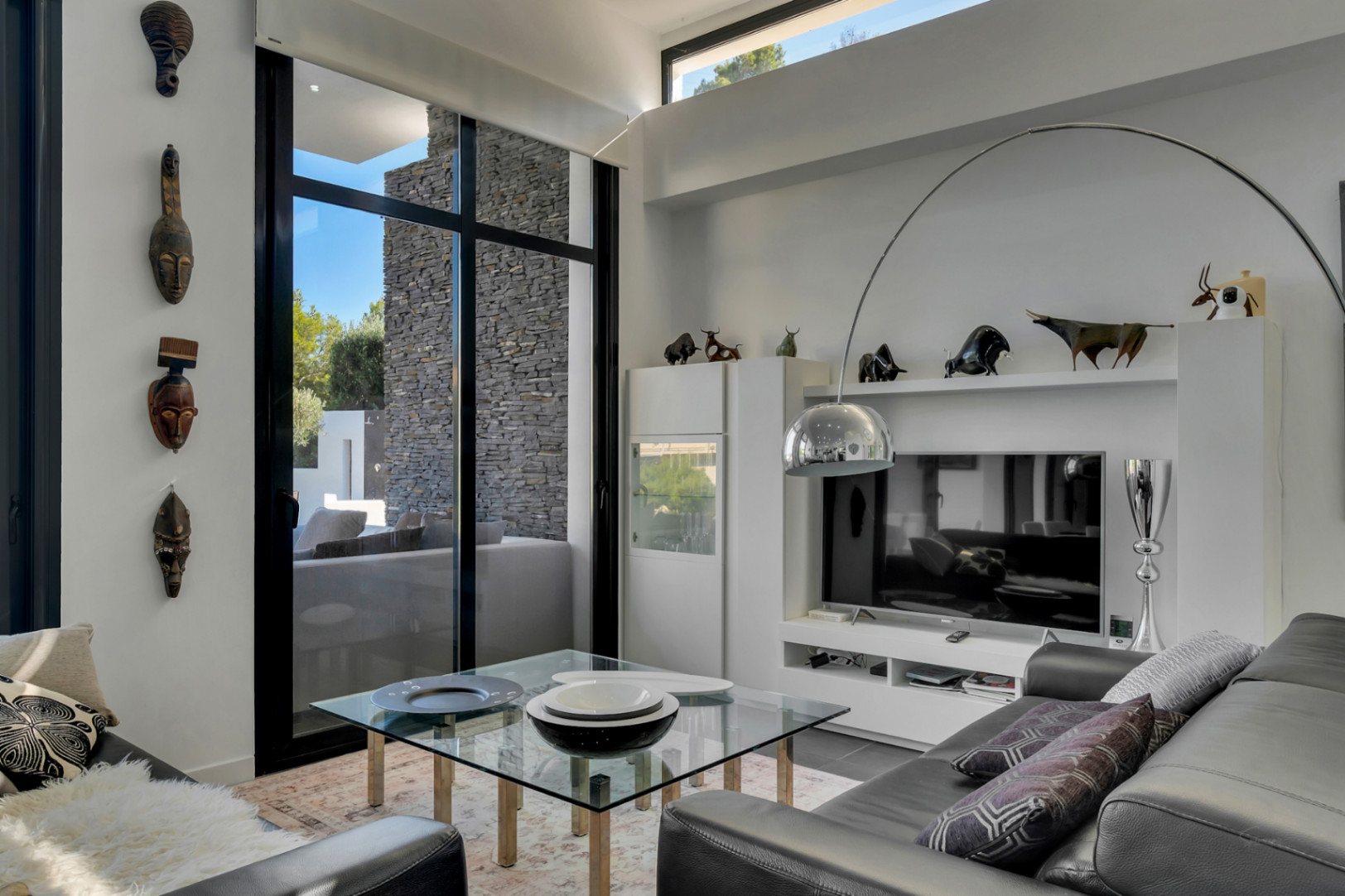 Moderne woonkamer met hoge plafonds, zwarte raamprofielen en etnische kunst. Voorzien van een chromen booglamp, glazen tafel en zicht op de natuurstenen buitenmuur.