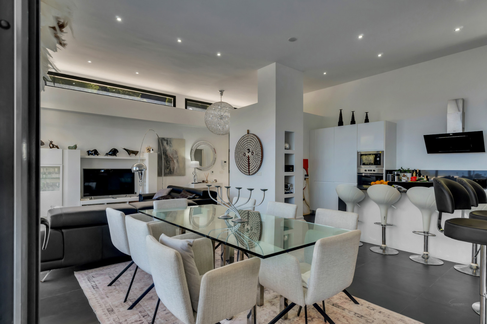 Open leefruimte met glazen eettafel, moderne keuken met ontbijtbar en hoge plafonds voorzien van inbouwspots en een kristallen kroonluchter.