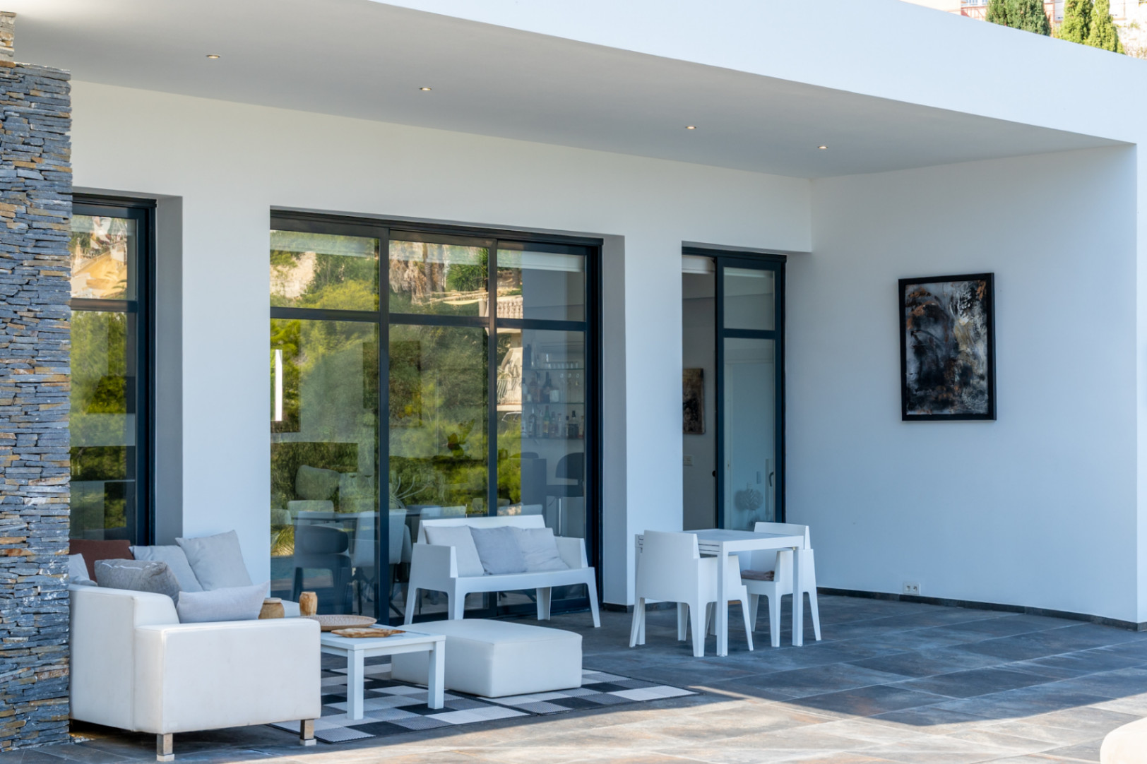 Overdekt terras met donkere tegels, wit designmeubilair en grote zwarte schuifpuien. Een natuurstenen kolom en inbouwspots zorgen voor een hoogwaardige afwerking.