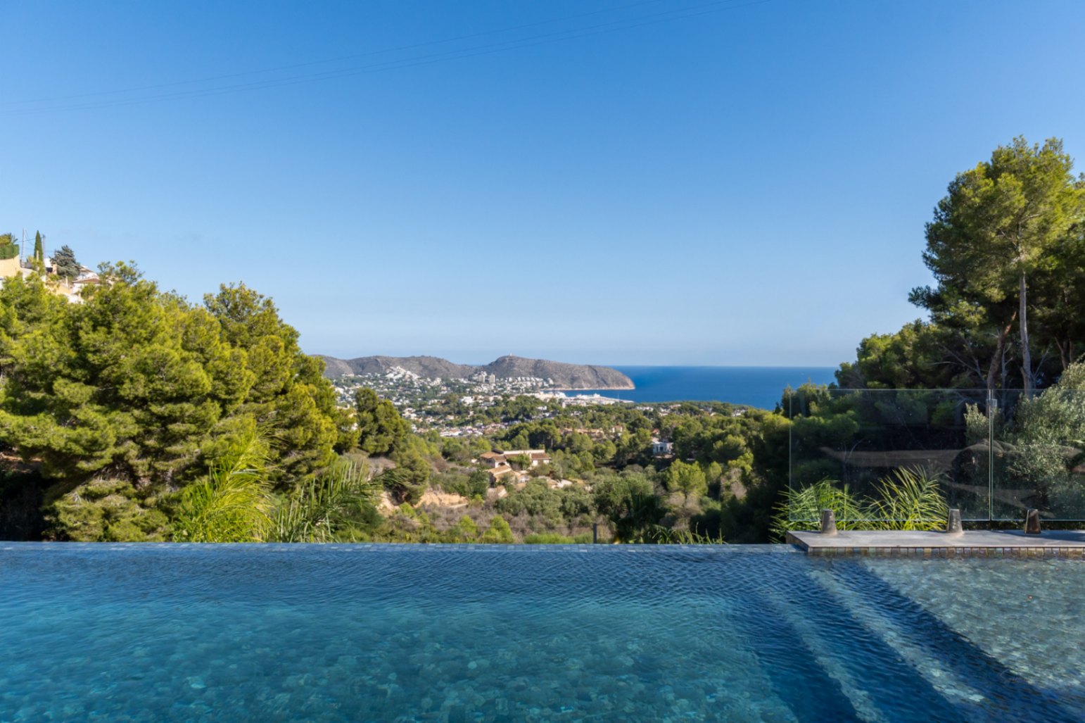 Panoramisch uitzicht op de Middellandse Zee en de kust van Moraira vanaf het overloopzwembad, omgeven door pijnbomen en lokale vegetatie.