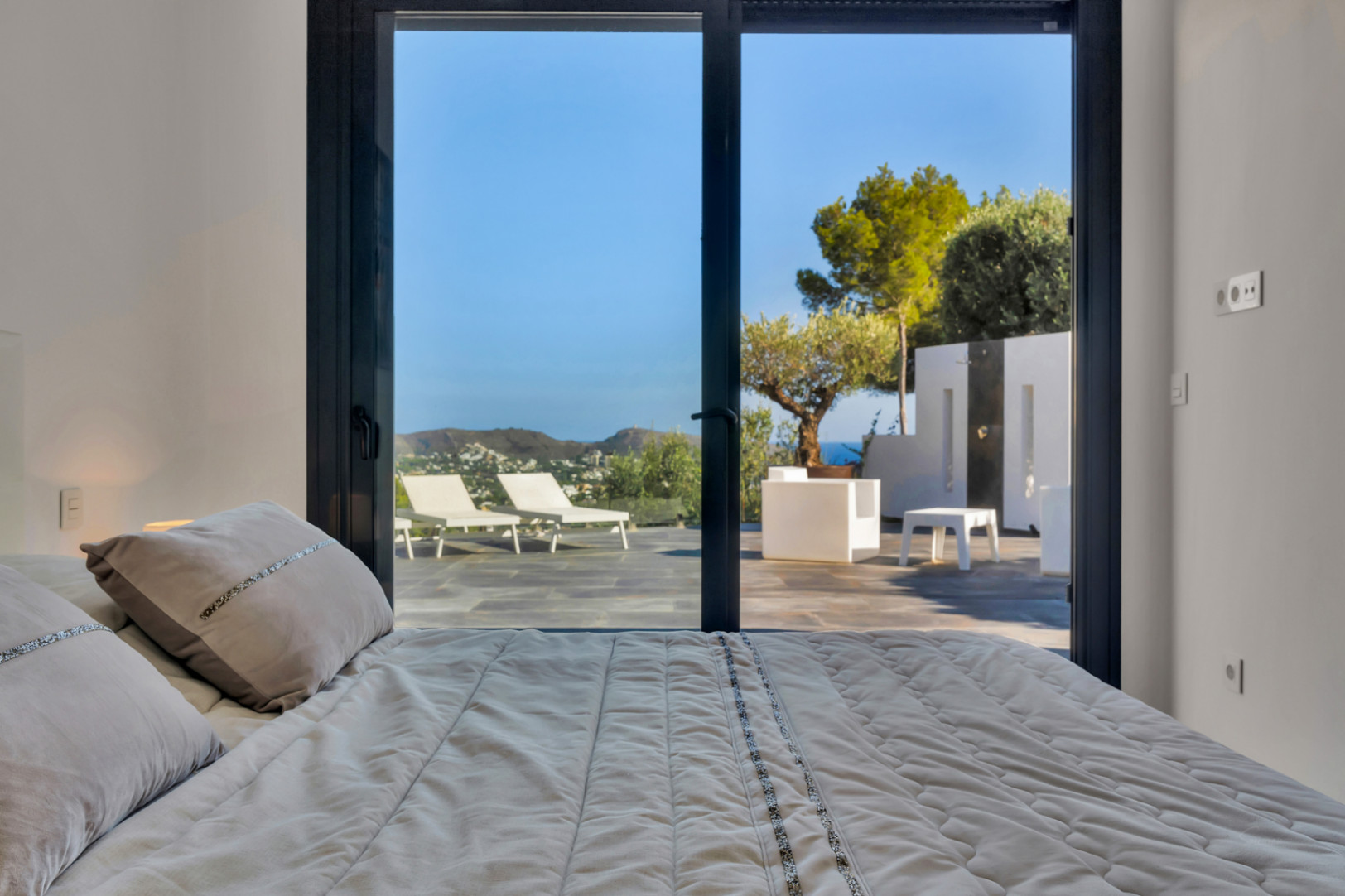 Slaapkamer met grote glazen schuifdeuren die toegang bieden tot een terras met natuursteen, witte ligbedden, een buitendouche en uitzicht op zee.