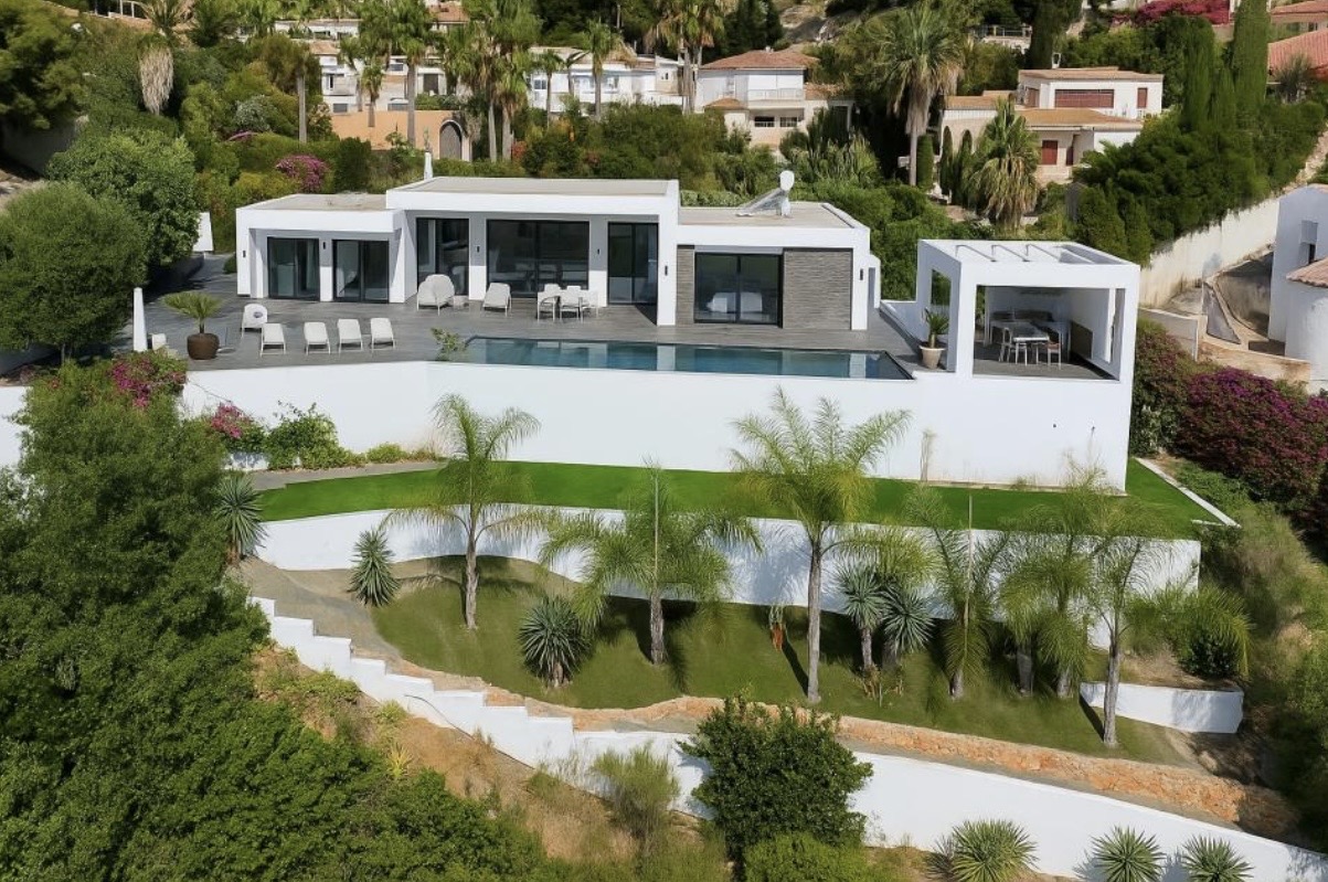 Verhoogd overzicht van de moderne witte villa met een strak zwembad, ruime zonneterrassen en een gelaagde tuin met palmbomen en gazons.