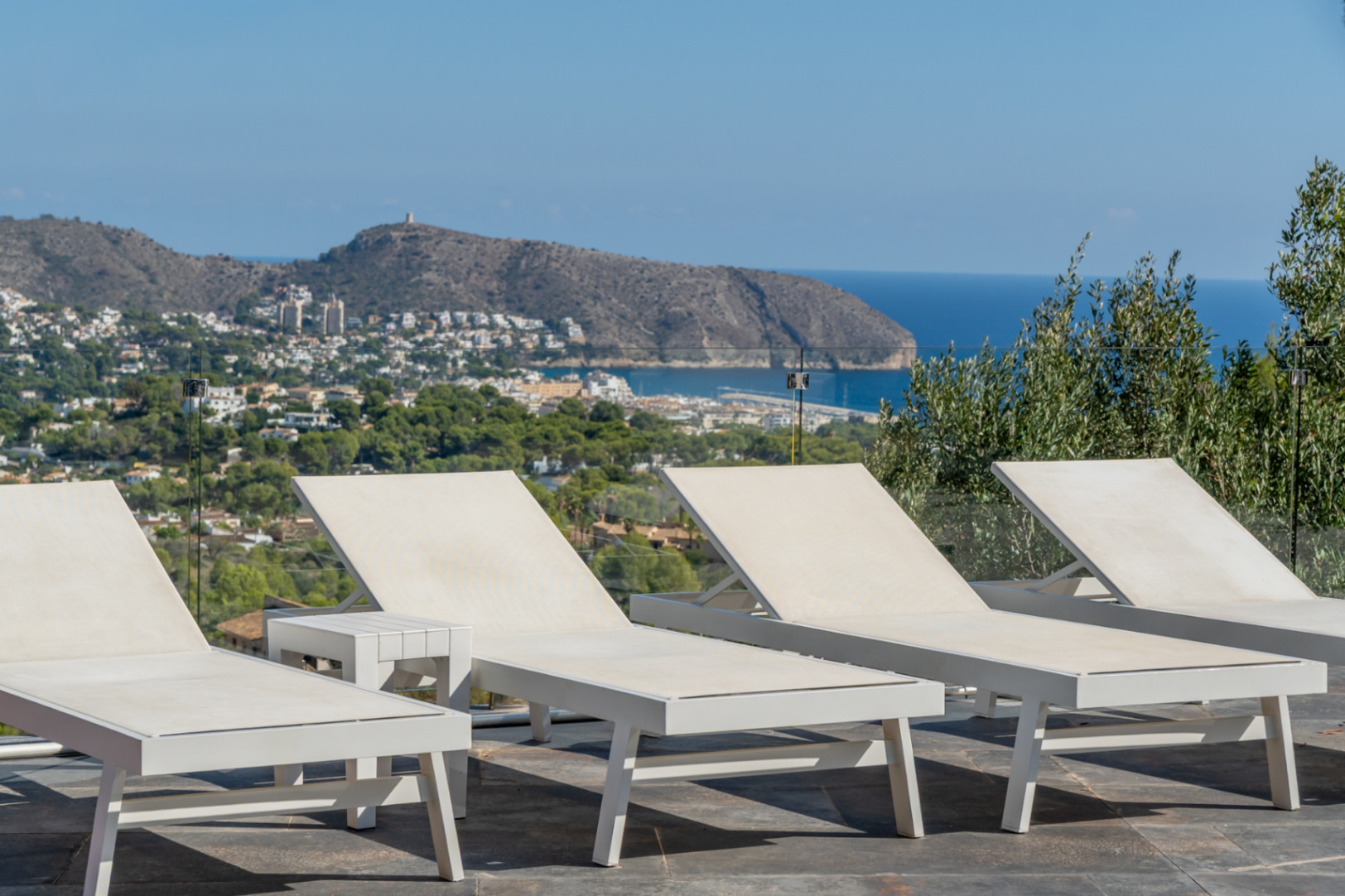Zonneterras met minimalistische witte ligbedden en glazen balustrades met panoramisch uitzicht op de Middellandse Zee en de kust van Moraira.