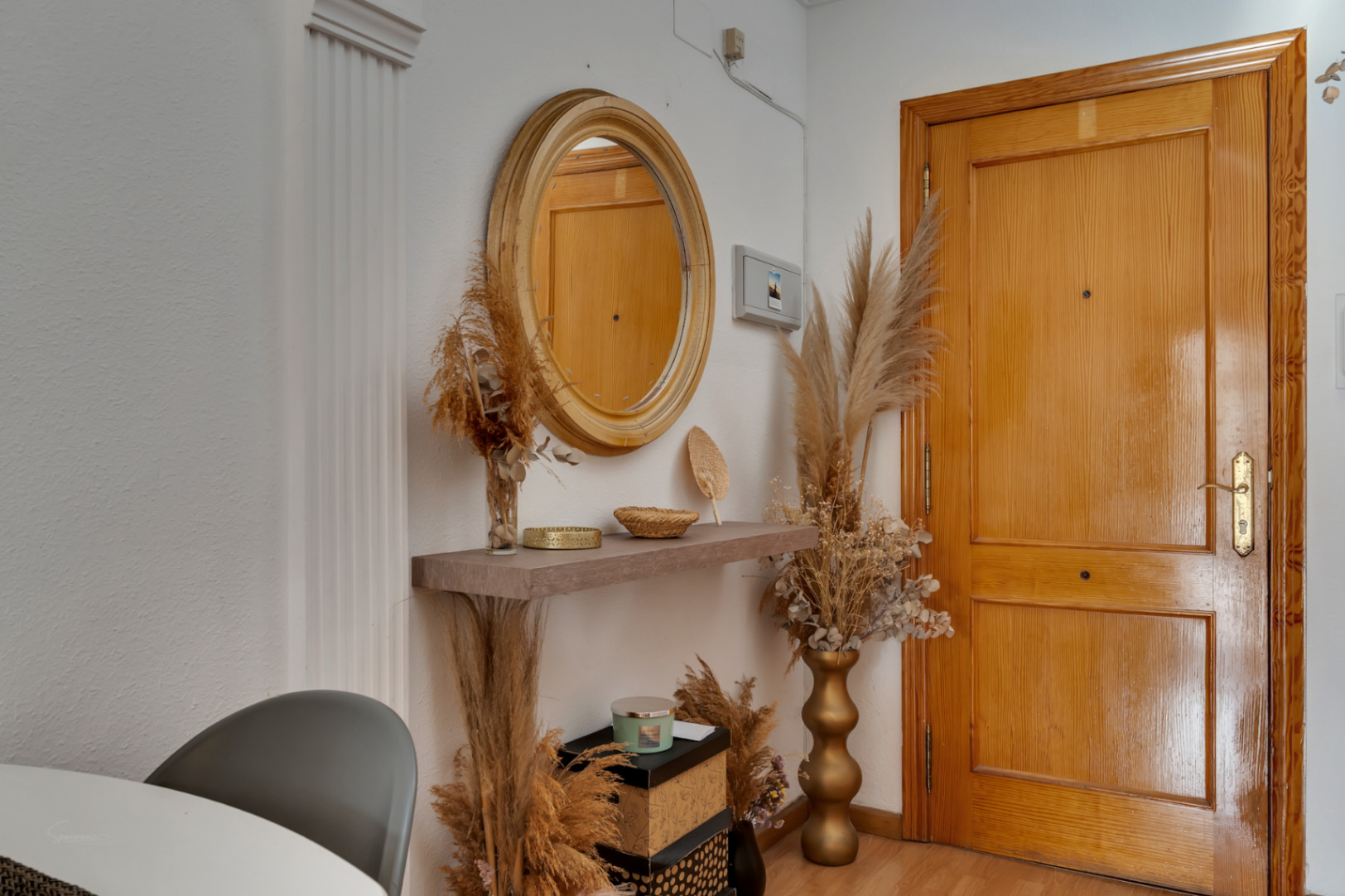 Entree met massief houten deur, ronde spiegel en zwevende plank gedecoreerd met droogbloemen en gouden accenten tegen witte wanden.