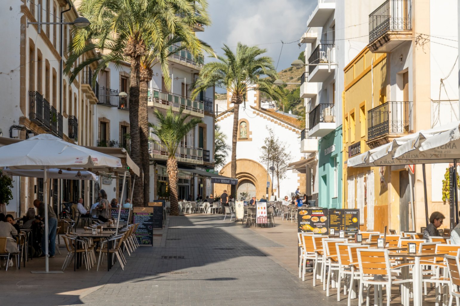Voetgangersstraat in het historische centrum van Jávea met terrassen, palmbomen en traditionele gevels die naar een historische kapel leiden.