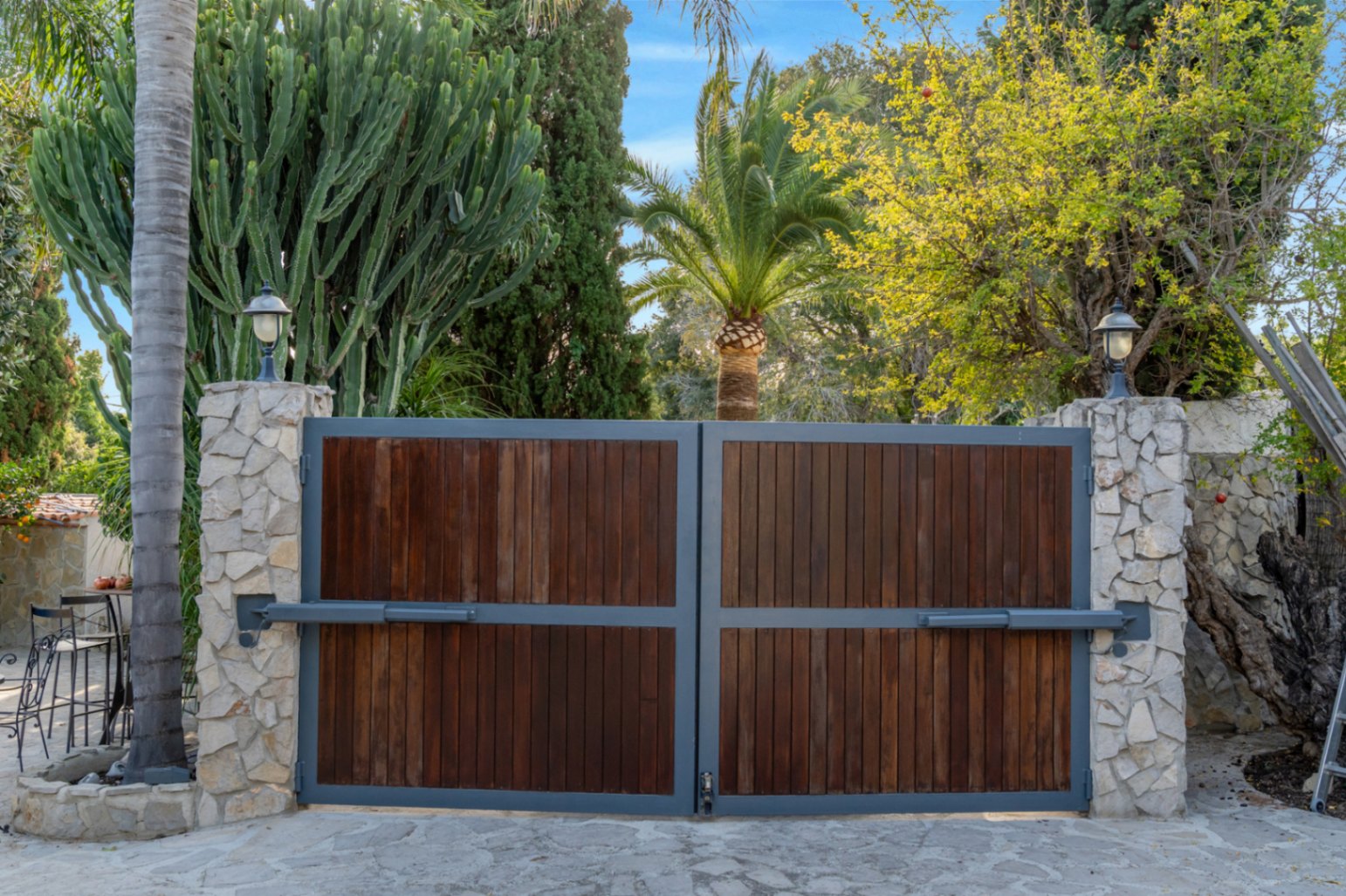 Automatische toegangspoort met houten panelen en een metalen frame, geflankeerd door natuurstenen pilaren en weelderige mediterrane beplanting.