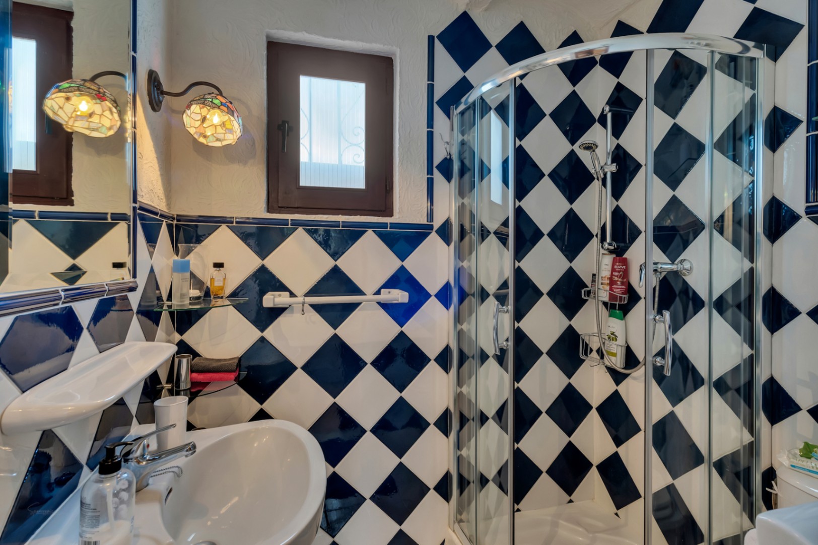 Badkamer met blauw-witte ruitvormige tegels, een halfronde glazen douchecabine en decoratieve wandlampen in Tiffany-stijl.