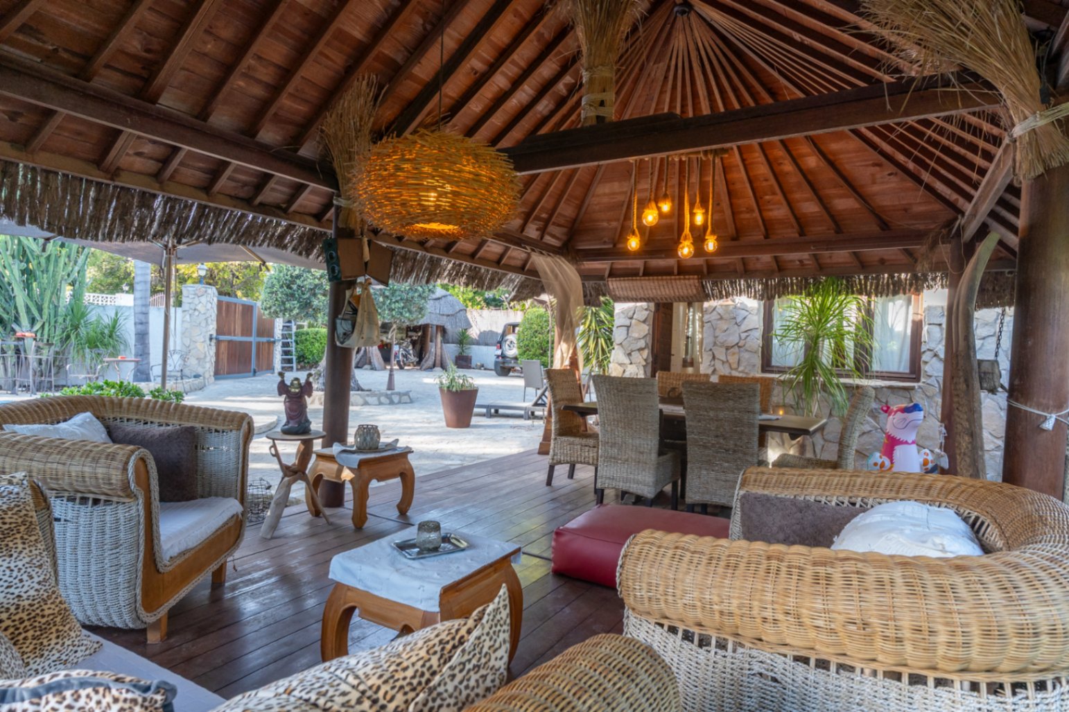 Balinese houten pergola met een open dakconstructie, rotan loungeset en een eethoek buiten op een donkerhouten vlonderterras.