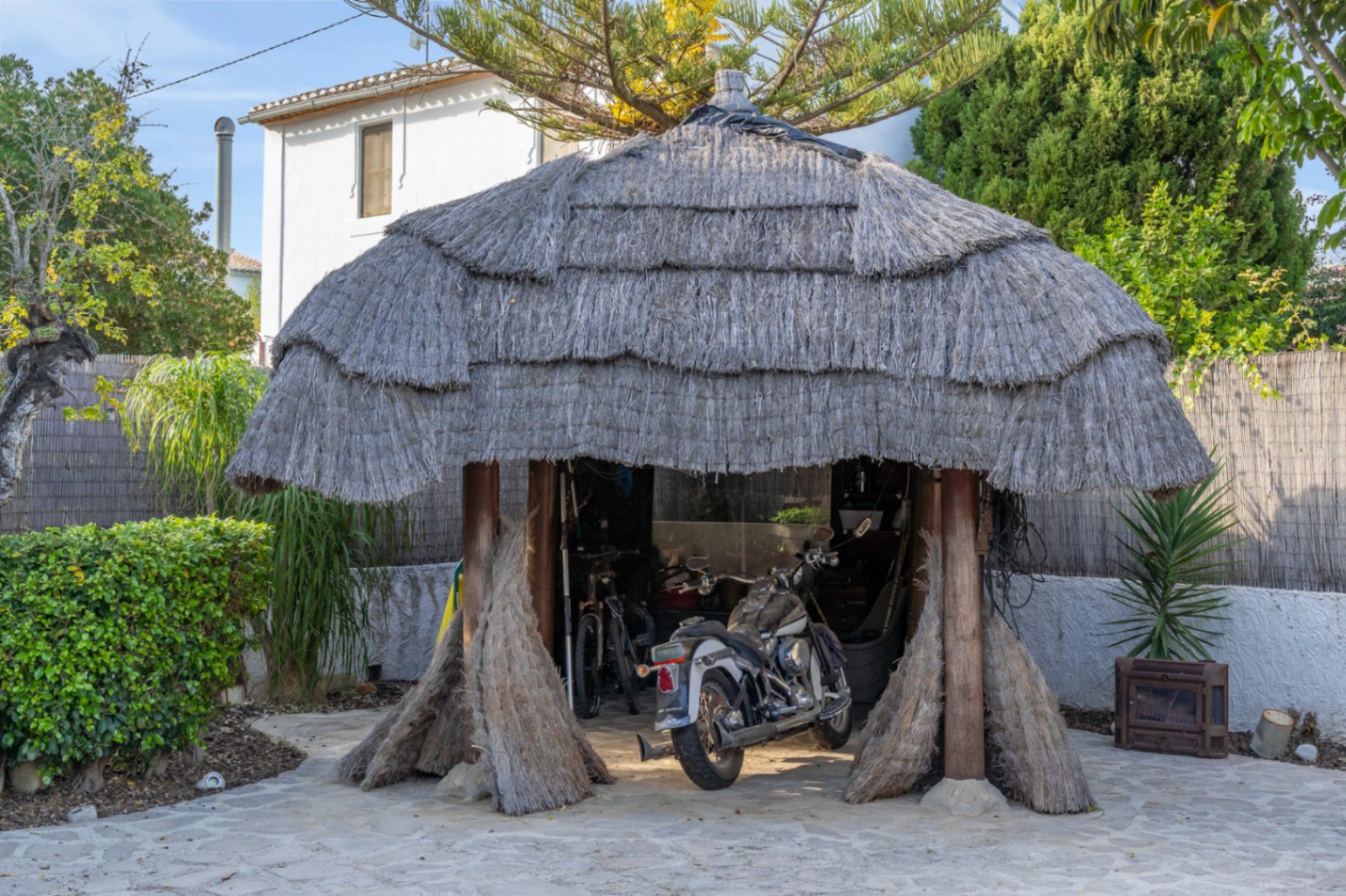 Balinese pergola met een dik rieten dak en houten steunbalken, momenteel in gebruik als overdekte parkeerplaats voor een motor en fietsen in de tuin.