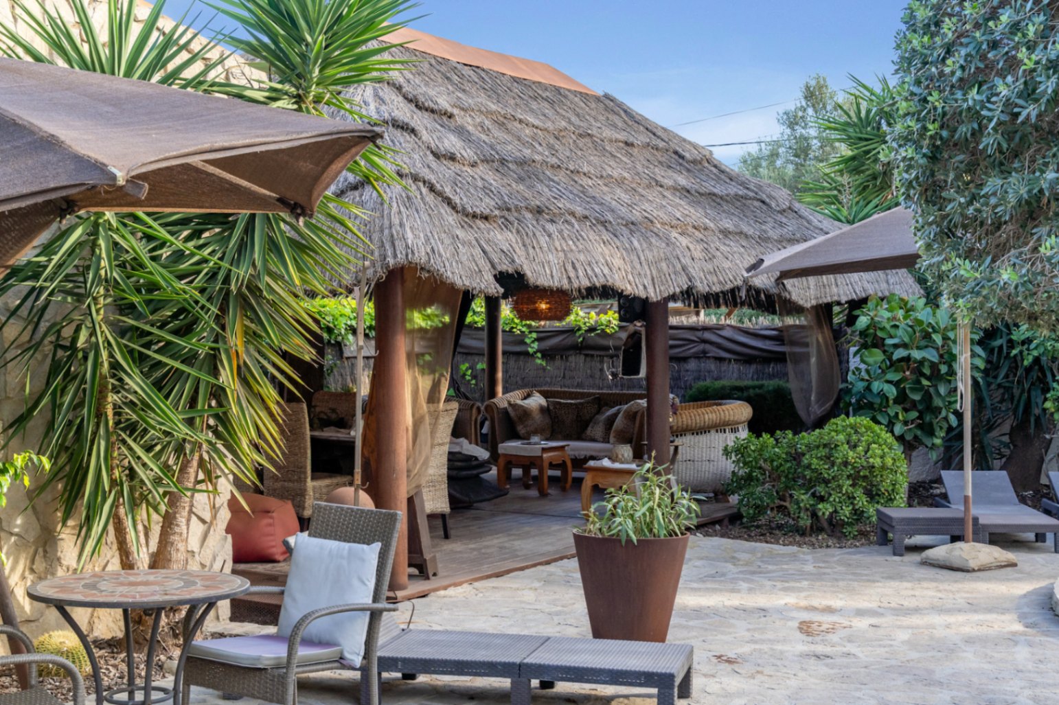 Balinese pergola met rieten dak en houten vlonder, gelegen op een natuurstenen terras omgeven door mediterrane beplanting en ligstoelen.