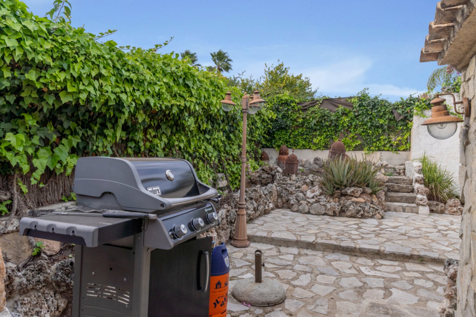Buitenbarbecueplaats met Weber-gasgrill, natuurstenen bestrating en weelderige klimopmuren in een rustieke mediterrane tuin.