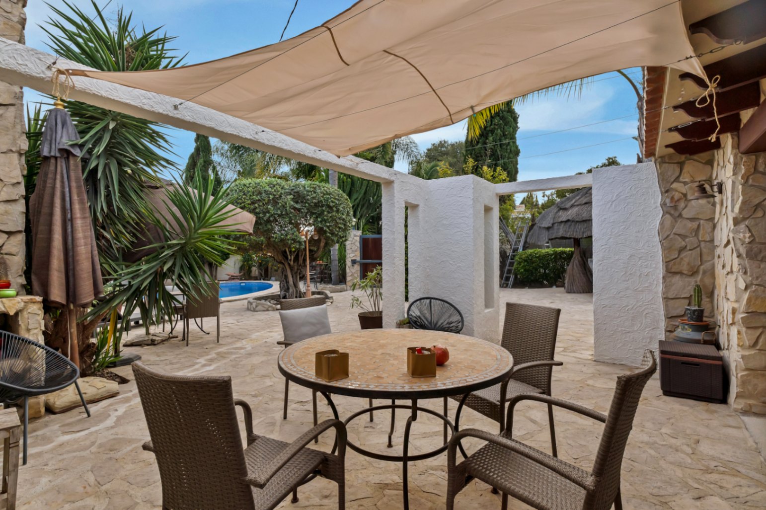 Buitenterras met natuurstenen vloer, een mozaïektafel onder een schaduwdoek en uitzicht op het zwembad en de mediterrane tuin.