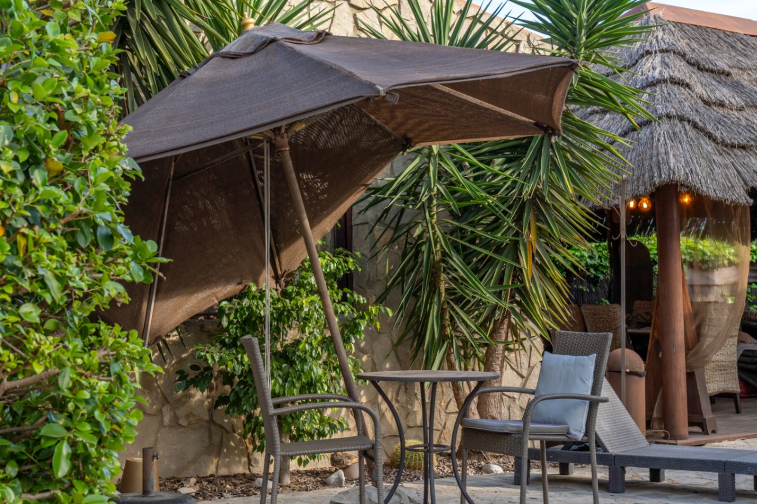 Buitenzitje met parasol, bistrotafel en rotan stoelen, gelegen bij een natuurstenen muur en een Balinese pergola met rieten dak.