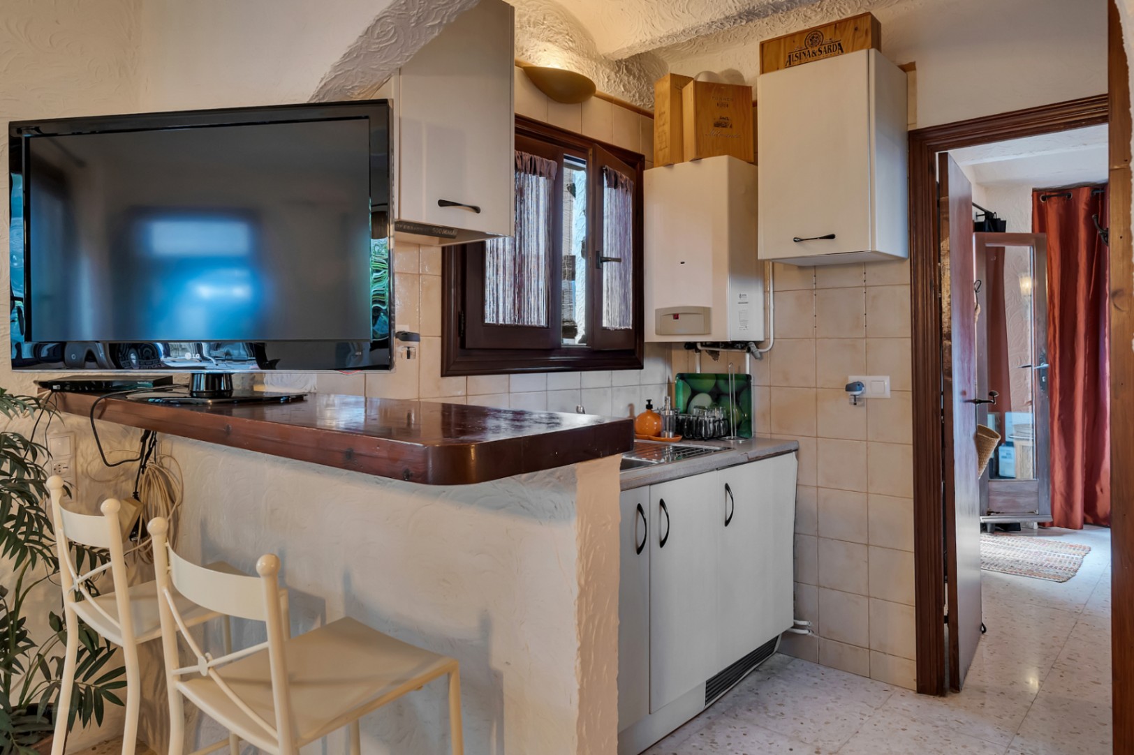 Compacte keuken met een donkerhouten ontbijtbar en witte krukken, voorzien van bovenkasten, een spoelbak en een aan de muur gemonteerde televisie.