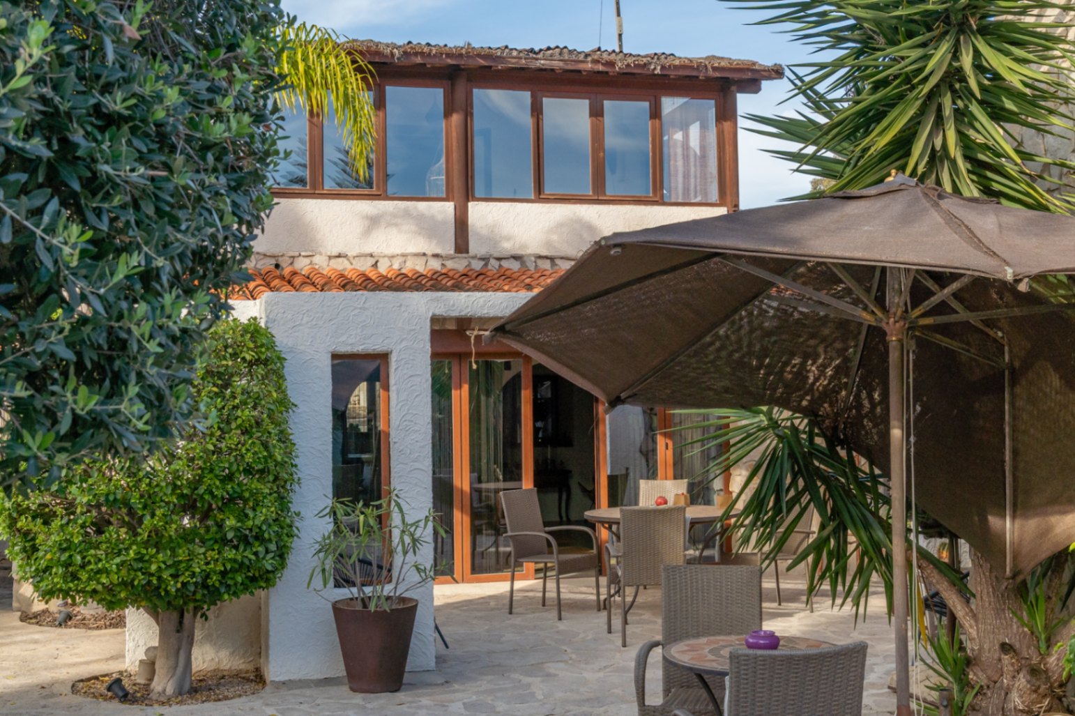 Exterieur van de villa met een beglaasd terras op de bovenverdieping, een stenen terras met buitenmeubilair en mediterrane beplanting.