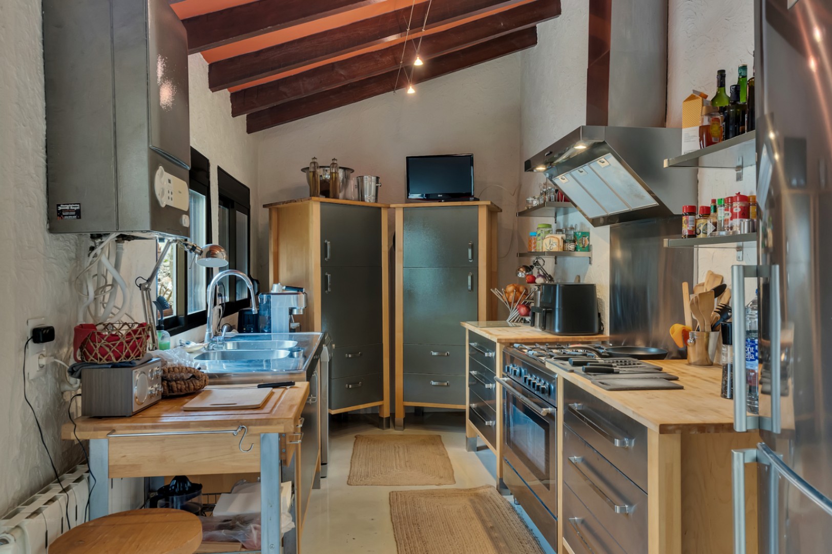 Industriële keuken met houten balkenplafond, massief houten werkbladen en roestvrijstalen apparatuur onder een schuin dak met terracotta tegels.