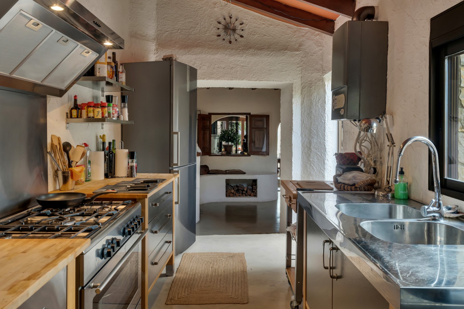 Moderne rustieke keuken met houten en roestvrijstalen werkbladen, witte structuurwanden en uitzicht op een woonruimte met een houtgestookte haard.