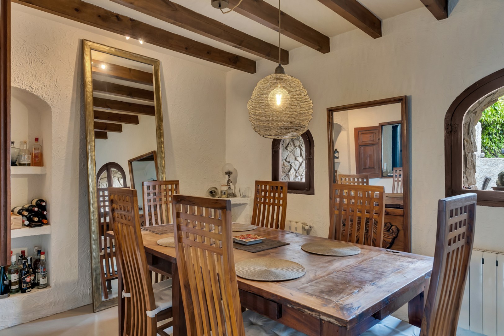 Rustieke eethoek met houten plafondbalken, een massief houten tafel en een hanglamp van natuurlijke vezels. Een grote vloerspiegel siert de witte muren met textuur.