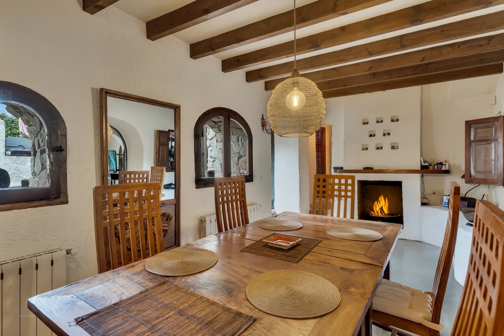 Rustieke eetkamer met houten balkenplafond, een massief houten tafel en een brandende open haard. Boogvensters en een geweven hanglamp versterken de mediterrane sfeer.