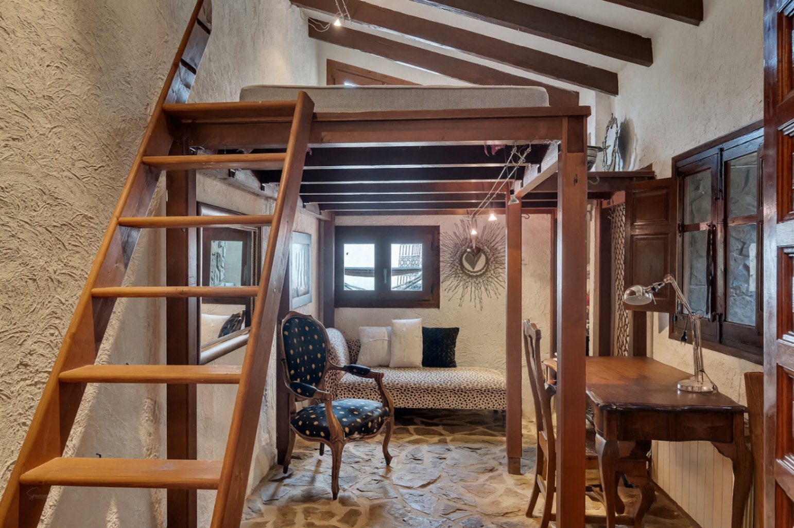 Rustieke slaapkamer met houten vide, zichtbare plafondbalken en een natuurstenen vloer. De ruimte is voorzien van een werkplek en zithoek onder de mezzanine.