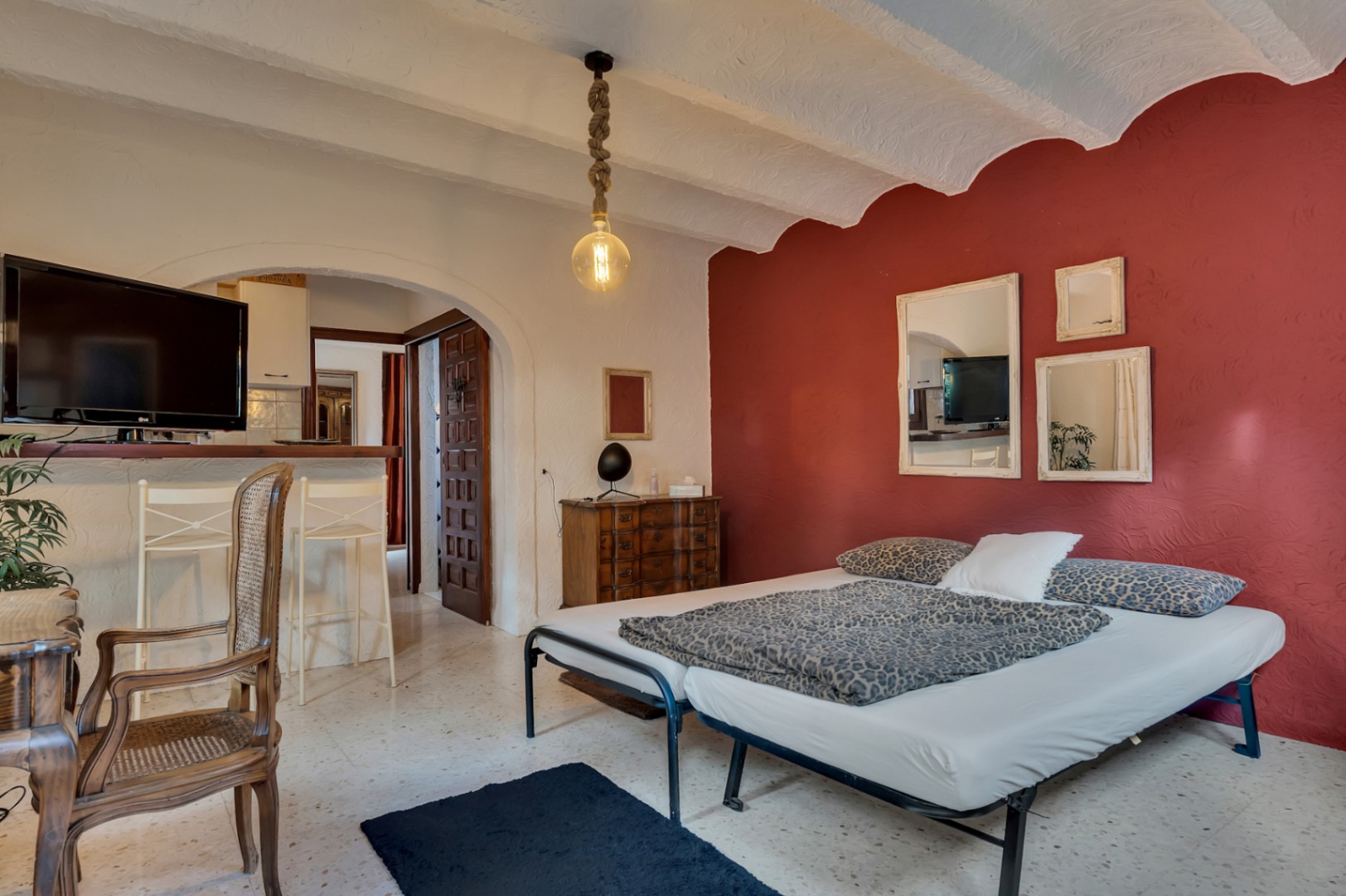 Slaapkamer met een rode accentmuur, wit gewelfd plafond en terrazzo vloer. Voorzien van een houten bureau, touwhanglamp en een bar die uitkijkt op de keuken.