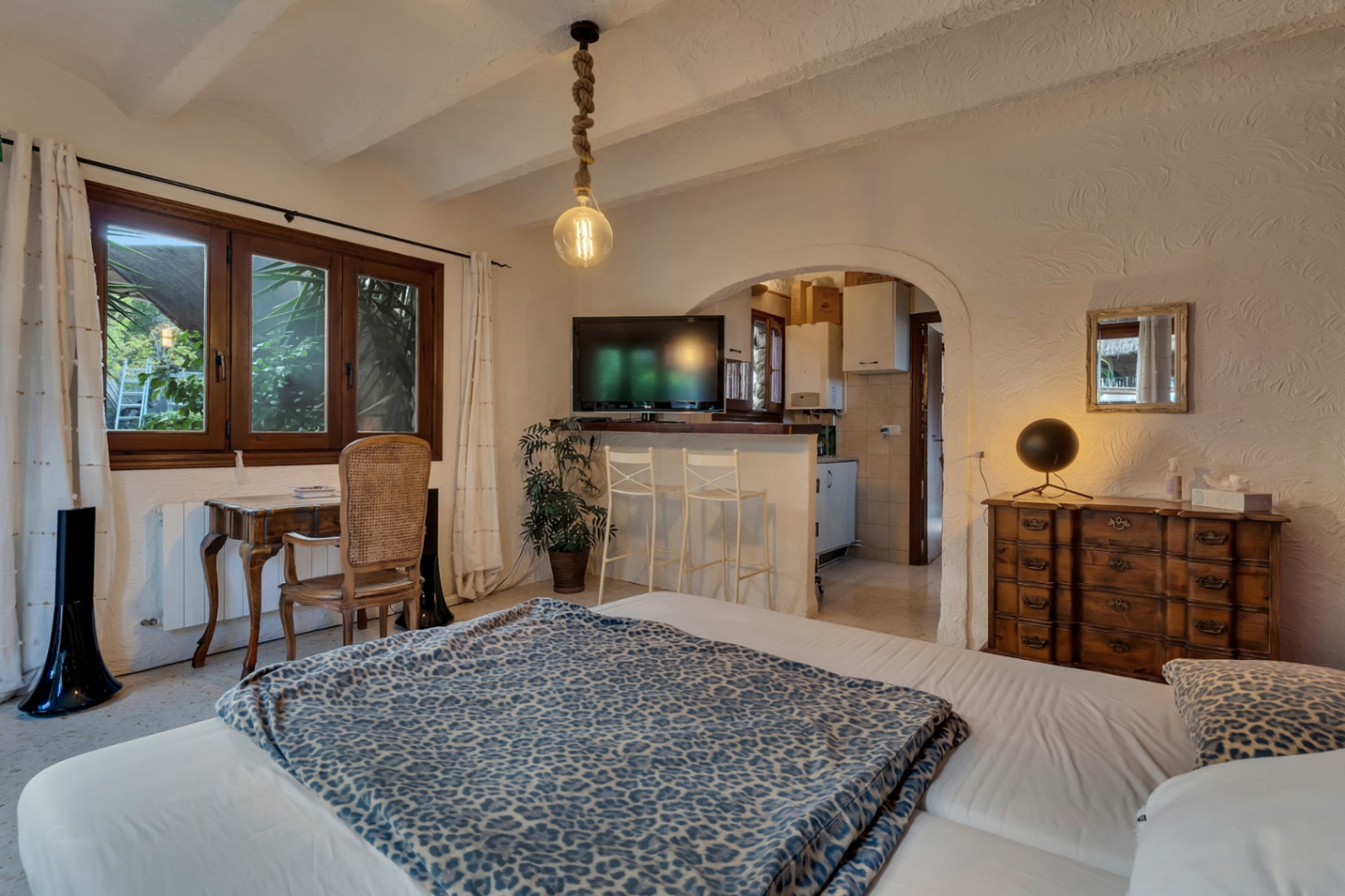 Slaapkamer met houten bureau, balkenplafond en een boogdoorgang naar een kitchenette met bar en krukken.