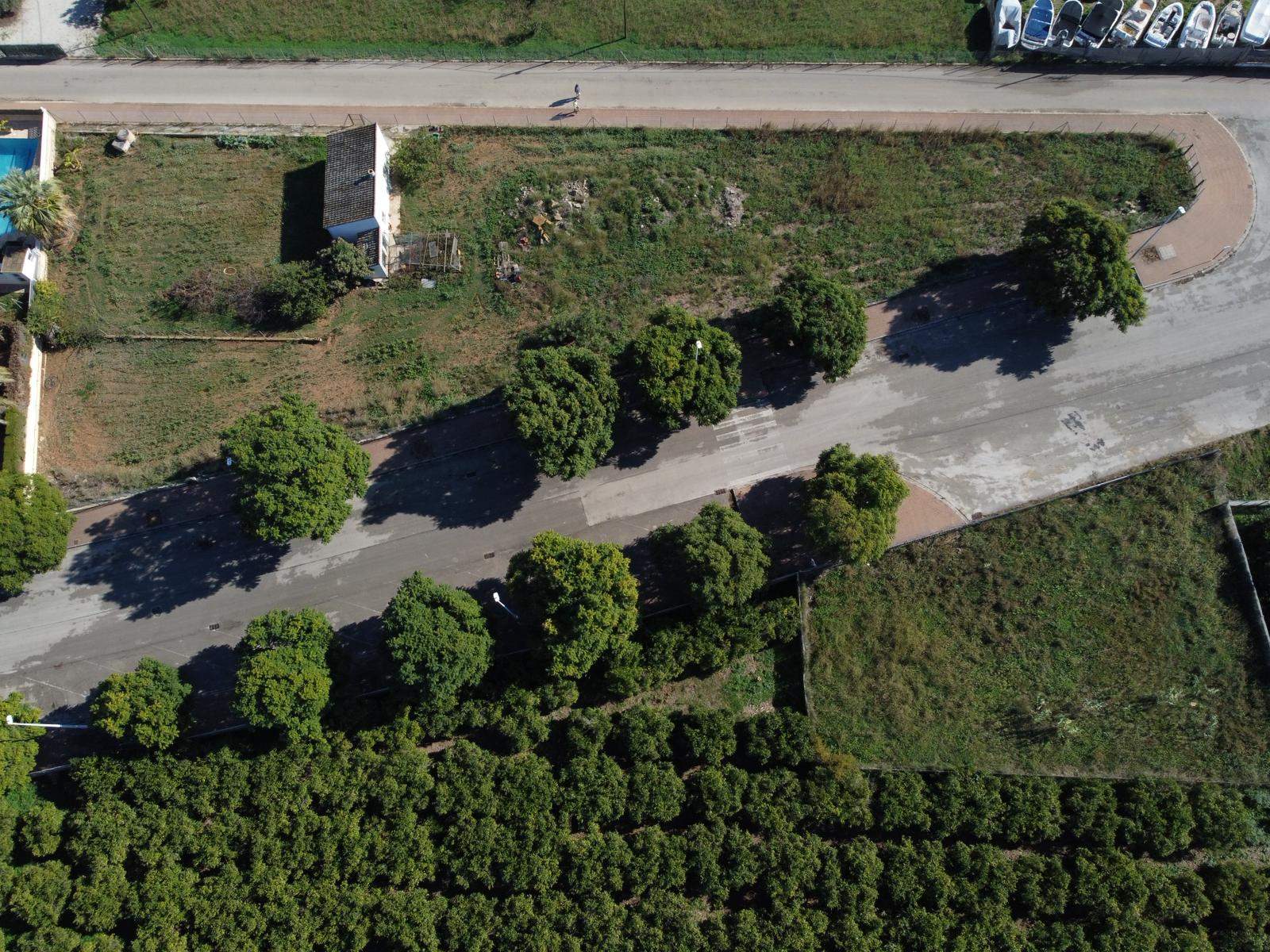 Bovenaanzicht van het stedelijke perceel met toegang via een geasfalteerde weg met bomen, een bestaand gebouwtje en de ligging naast landbouwgrond.