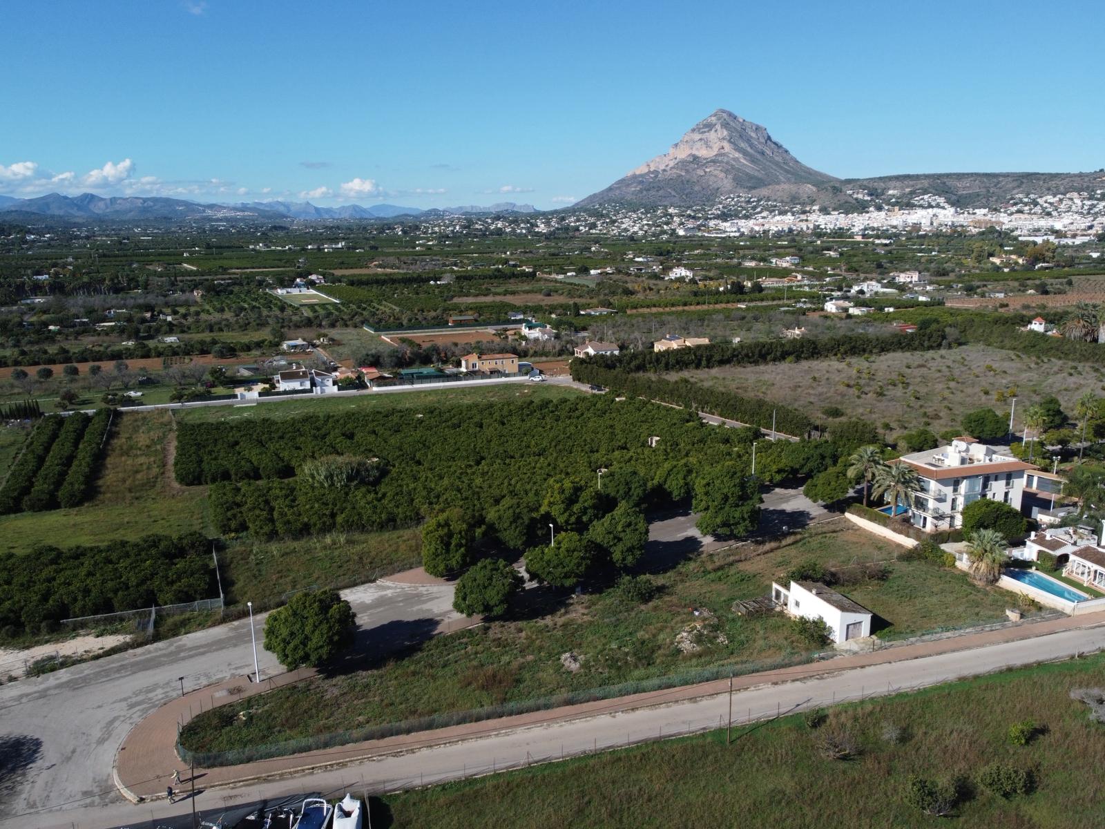 Luchtfoto van het bouwperceel met verharde toegangsweg, grenzend aan groene landbouwgronden met de iconische Montgó-berg op de achtergrond.