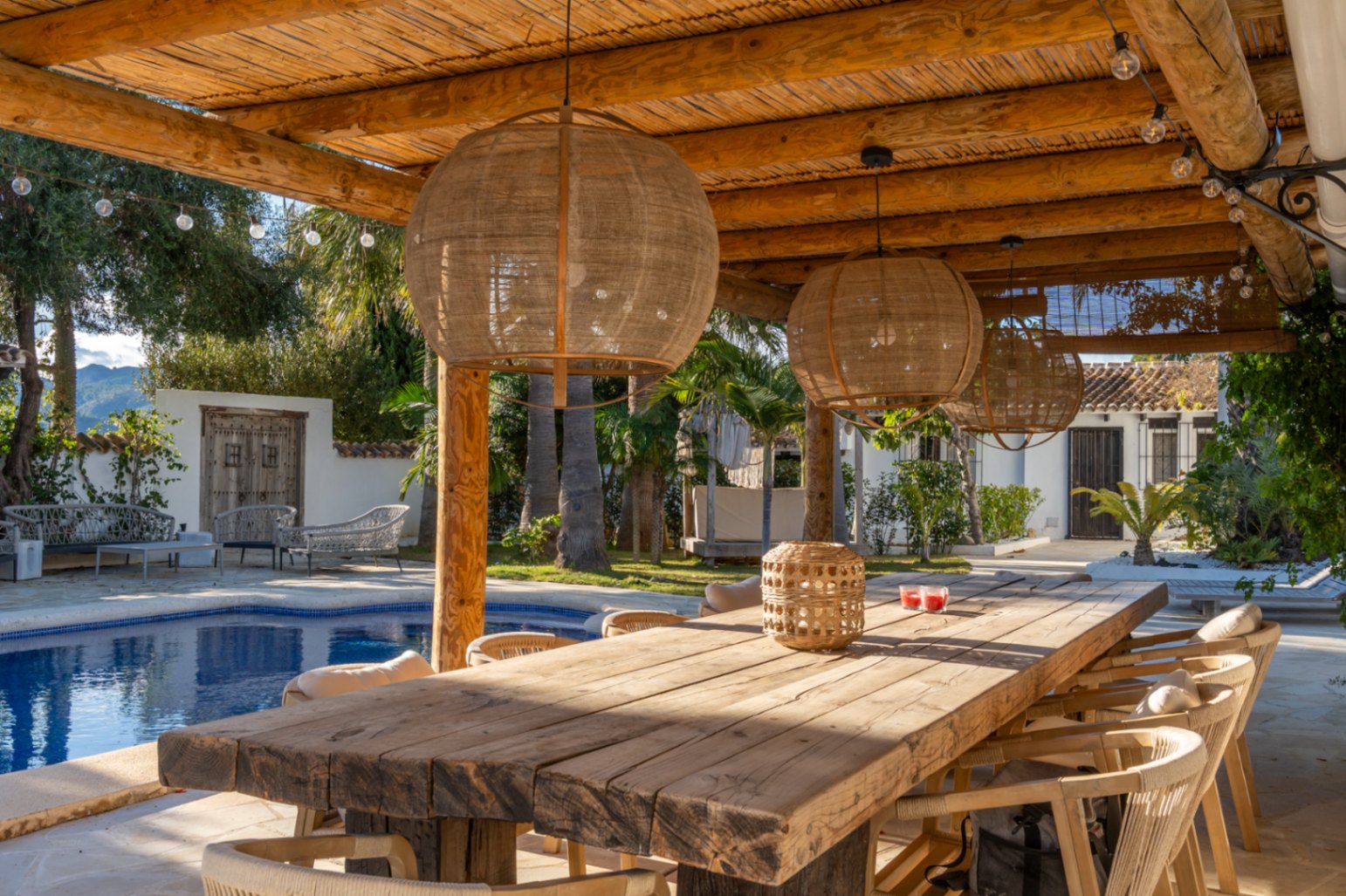 Buitenruimte met eethoek onder een houten pergola met rieten dak, voorzien van een rustieke tafel, natuurlijke hanglampen en uitzicht op het zwembad en de tuin.