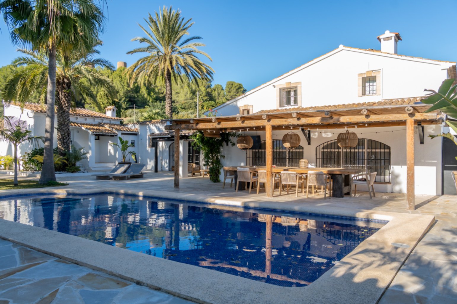 Buitenruimte met rechthoekig zwembad en houten pergola. Het terras is voorzien van een grote eettafel, rieten hanglampen en een authentieke mediterrane uitstraling.