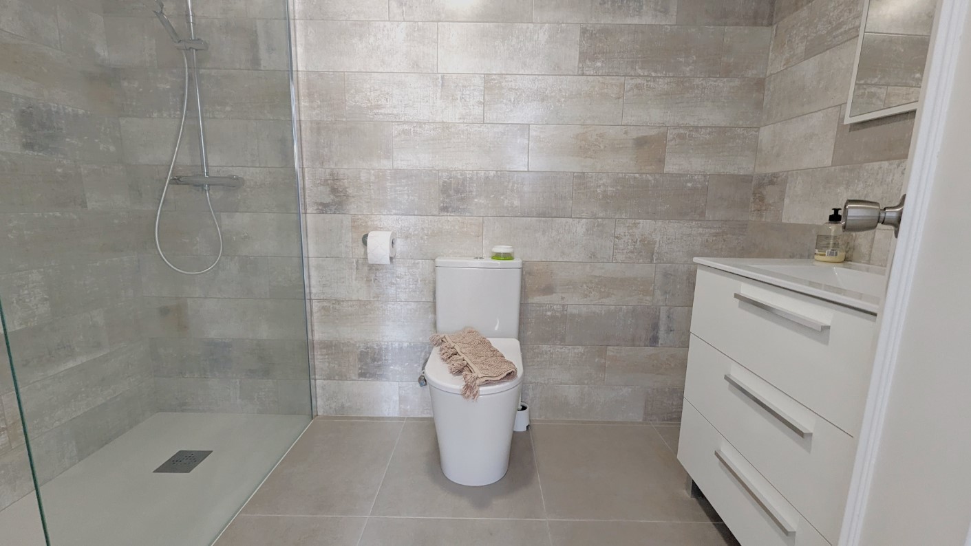 Moderne badkamer met inloopdouche, glazen wand, grijze getextureerde wandtegels en een wit badkamermeubel met lades en wastafel.