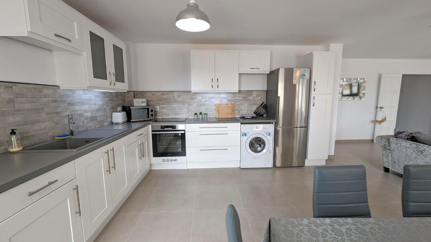 Moderne open keuken met witte kasten in shaker-stijl, grijze achterwand met steeneffect en geïntegreerde roestvrijstalen apparatuur waaronder een wasmachine.