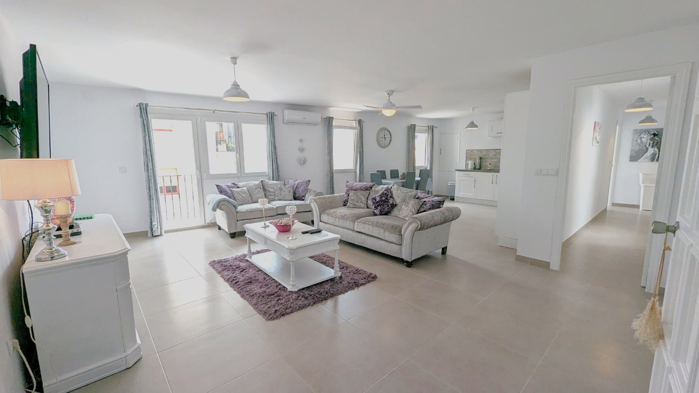 Open woonkamer met grijze banken, paarse accenten en een moderne witte keuken. De ruimte is voorzien van een lichte tegelvloer, plafondventilator en toegang tot de balkons.