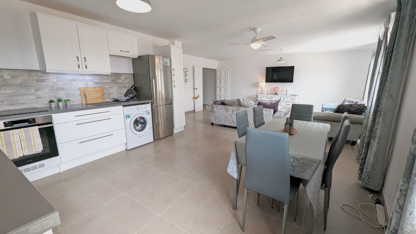 Open woonruimte met een moderne witte keuken inclusief inbouwapparatuur, een eethoek met grijze stoelen en een zithoek met plafondventilator en lichte tegelvloer.
