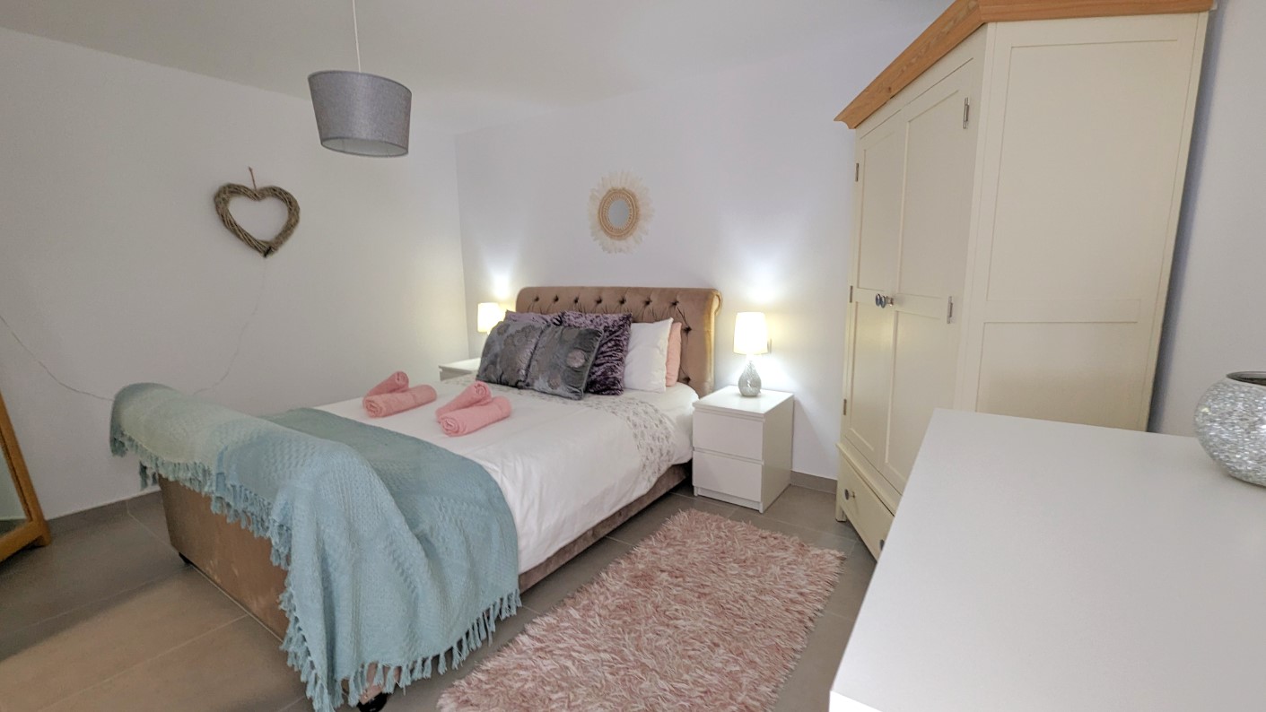 Slaapkamer met een tweepersoonsbed met gecapitonneerd hoofdbord, witte nachtkastjes en een crèmekleurige kledingkast. Voorzien van een tegelvloer en neutrale wanden.