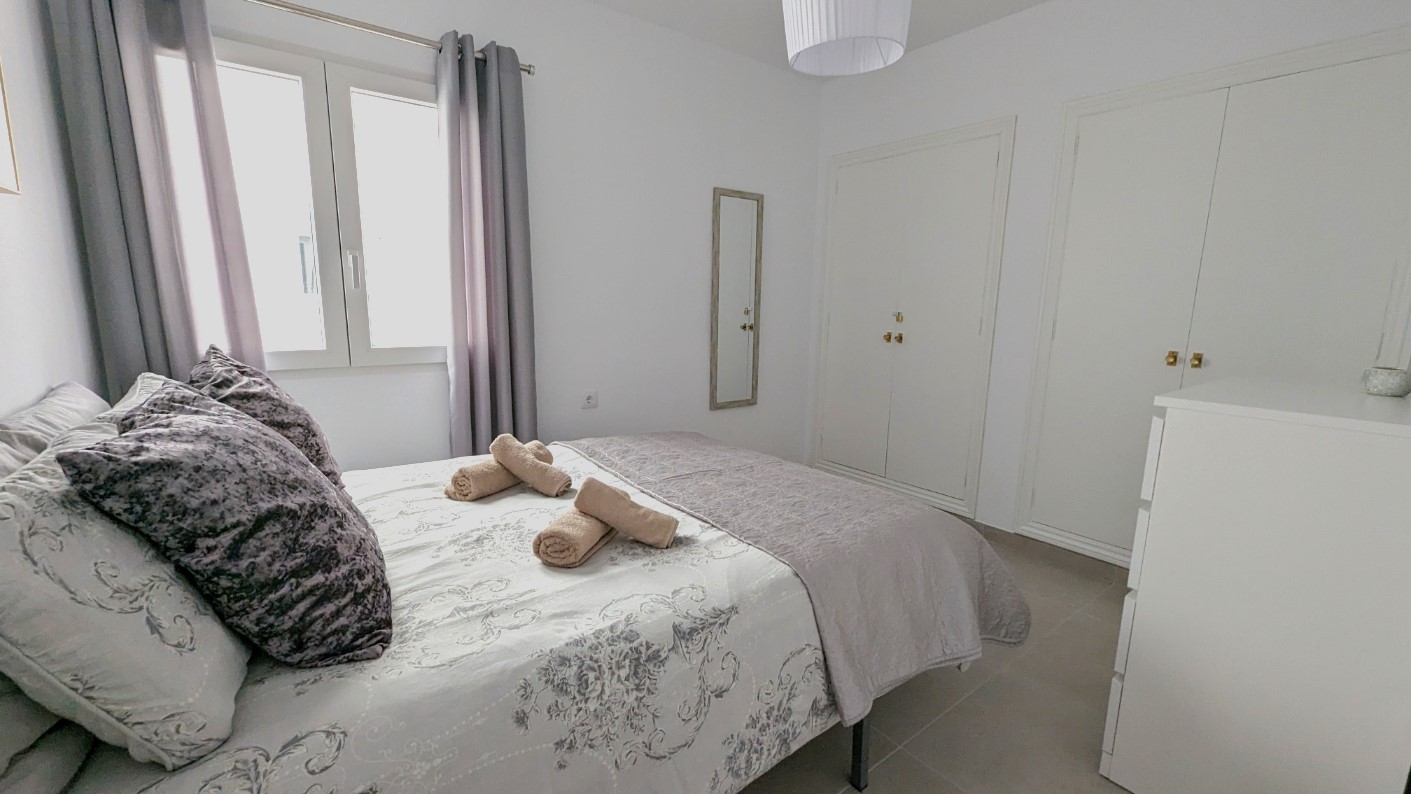 Tweepersoonsslaapkamer met witte inbouwkasten, een wandspiegel en een tegelvloer. De kamer heeft veel natuurlijk lichtinval en een afwerking in neutrale grijstinten.