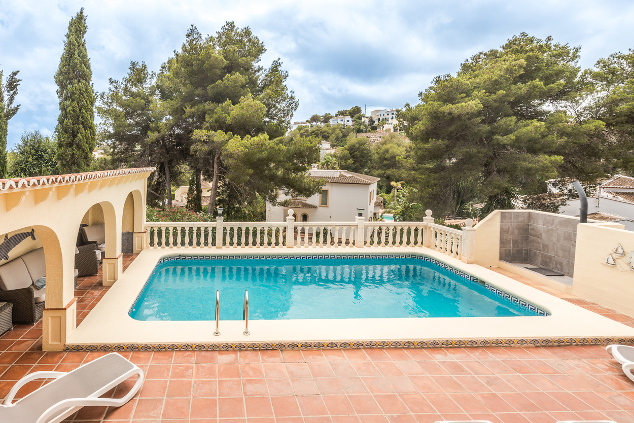 Buitenterras met een zwembad van 8x4m, witte balustrade en een overdekte naya met bogen, omgeven door volgroeide pijnbomen en mediterrane beplanting.
