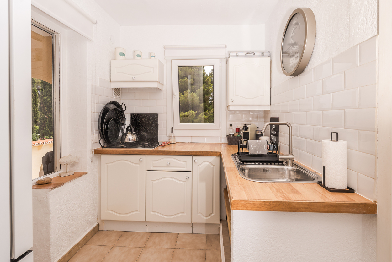 Compacte keuken met witte kasten, houten werkbladen en een witte metrotegel achterwand. Voorzien van gaskookplaat, roestvrijstalen spoelbak en uitzicht op de tuin.