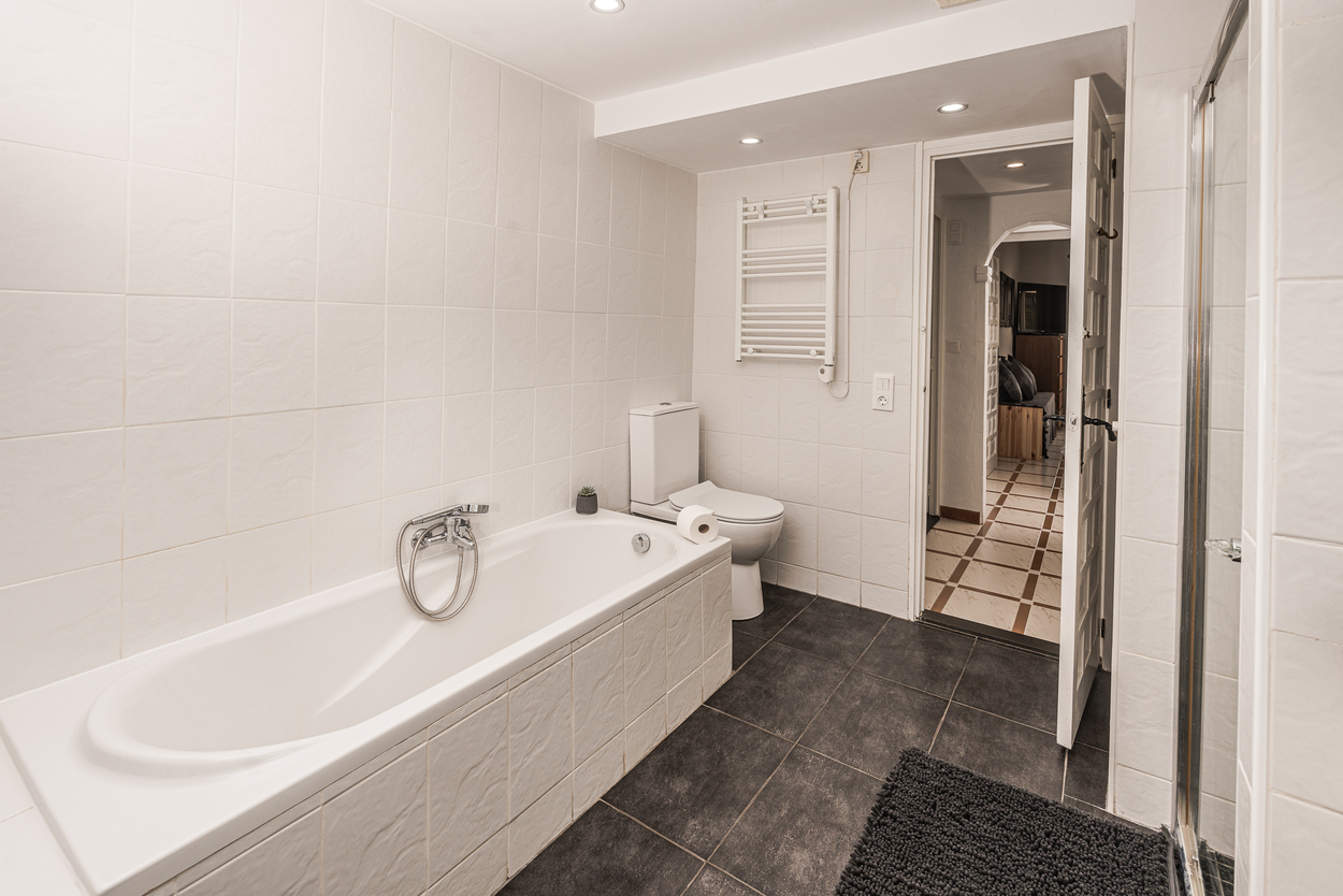 Functionele badkamer met een wit betegeld ligbad, donkere vloertegels, een handdoekradiator en inbouwspots in het plafond.