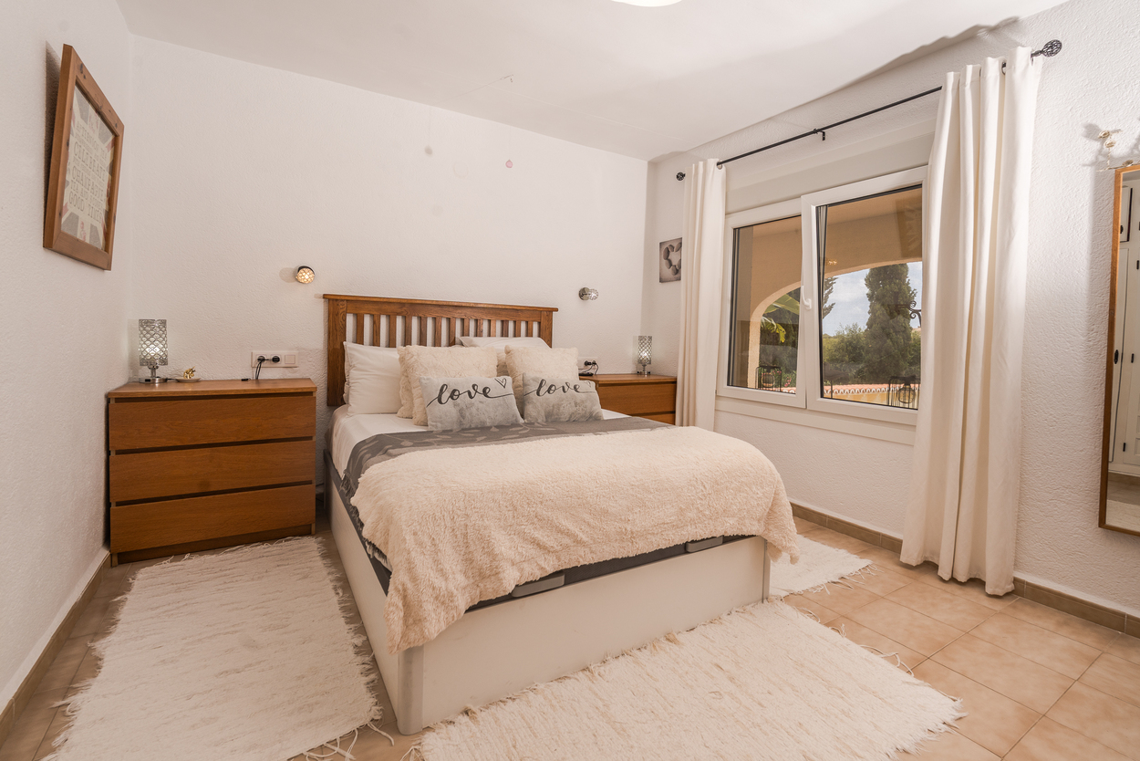 Lichte slaapkamer met houten hoofdbord, dubbele beglazing en tegelvloer. Voorzien van bijpassende houten ladekasten en uitzicht op de omliggende tuin.