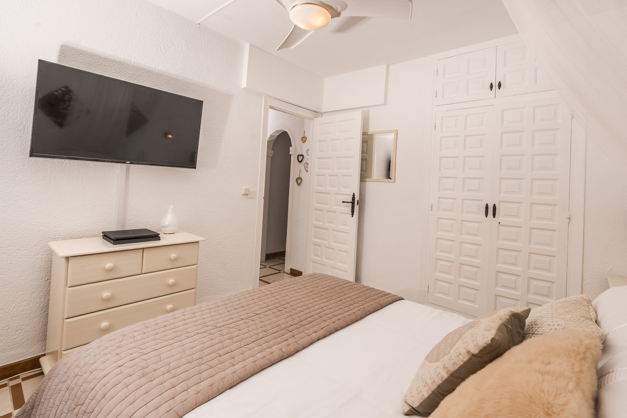 Lichte slaapkamer met witte ingebouwde kasten, een wandtelevisie en een plafondventilator. Voorzien van een bijpassende ladekast en tegelvloer.