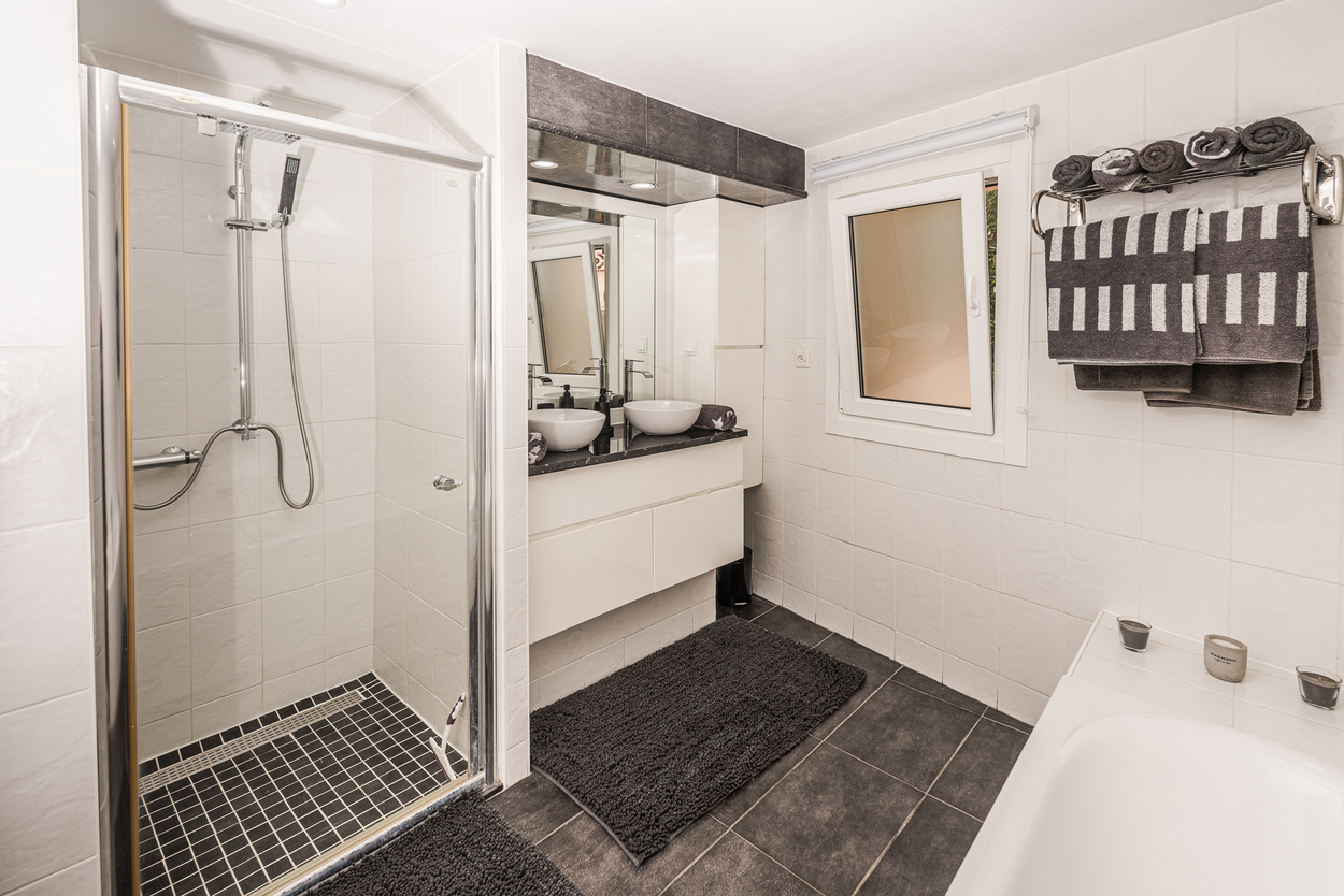 Moderne badkamer met glazen douchecabine, dubbele wastafel op een donker blad en contrasterende antracietkleurige vloertegels.