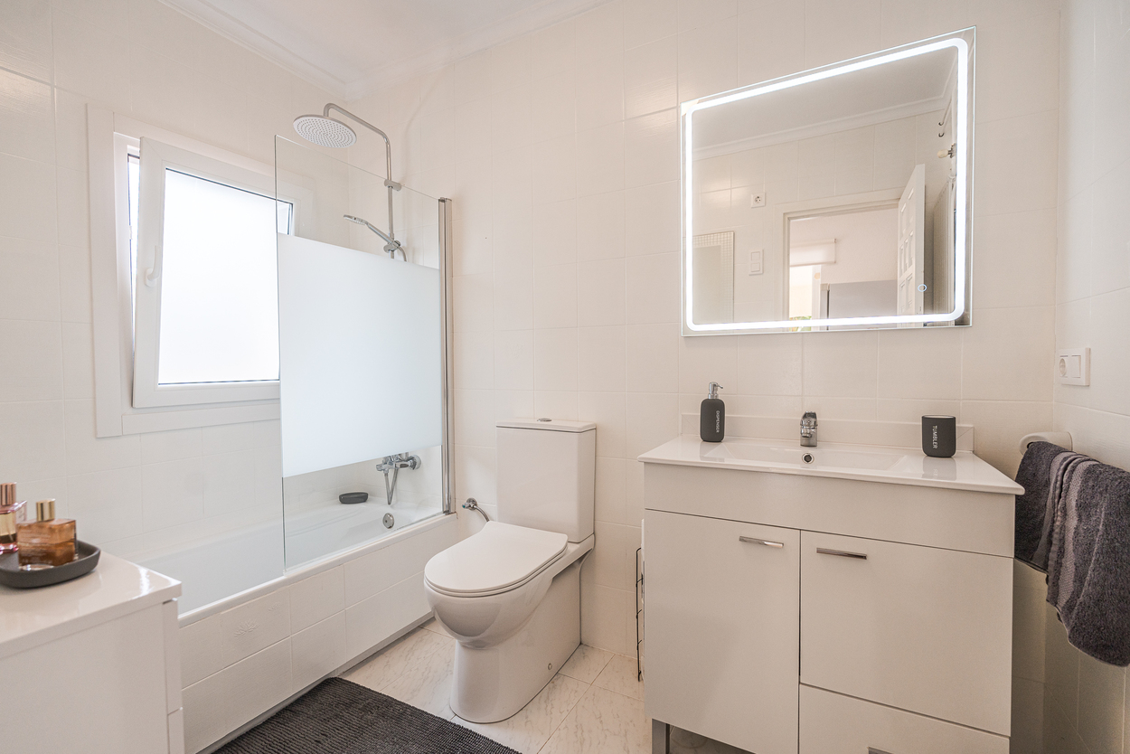Moderne badkamer met witte betegeling, wastafelmeubel met LED-verlichte spiegel en een ligbad met glazen douchescherm en regendouche.
