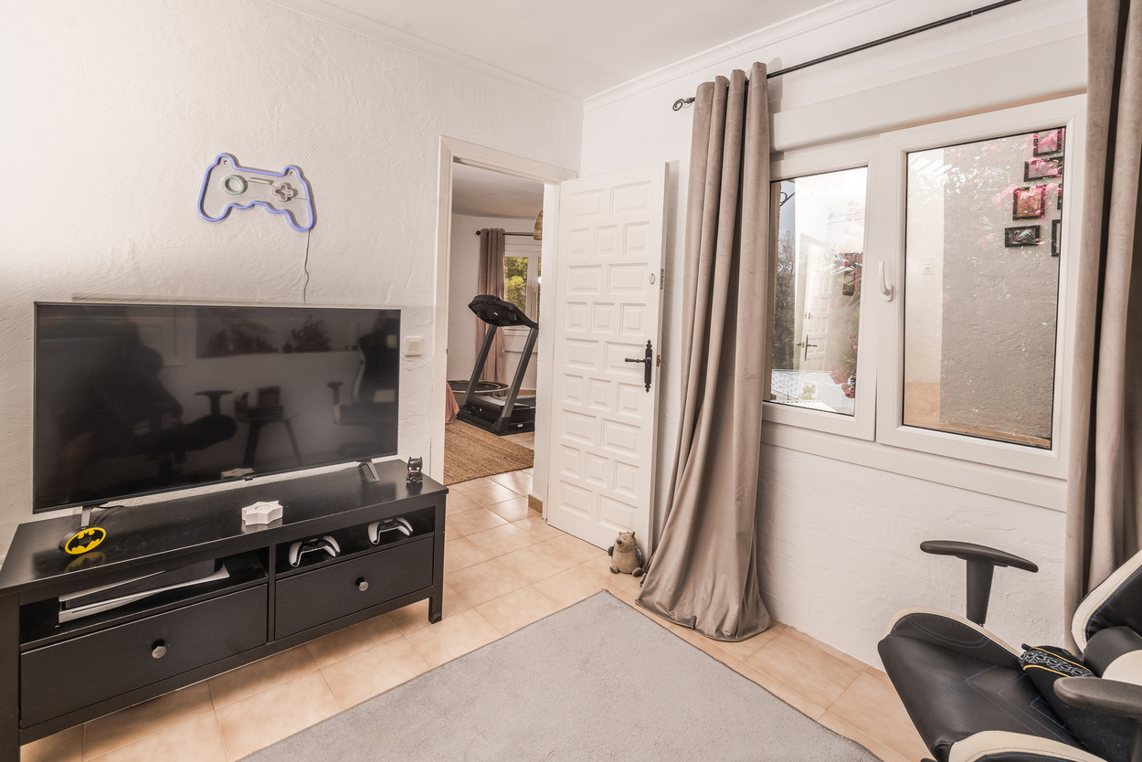 Multimediakamer met gaming-opstelling, flatscreen-tv en tegelvloer, met uitzicht op een aangrenzende fitnessruimte met loopband.