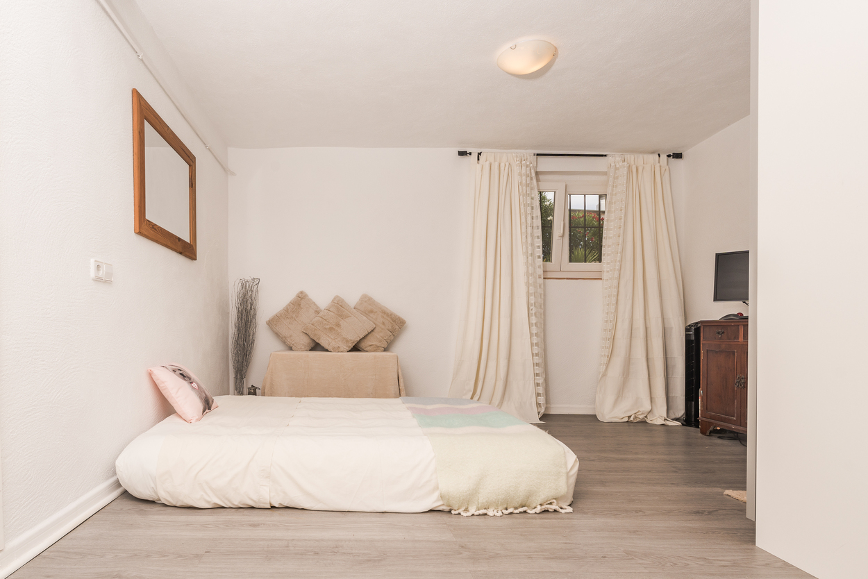 Slaapkamer met grijze laminaatvloer in houtlook, witte muren en een laag bed. Voorzien van een donkerhouten kast, spiegel met houten lijst en raam met gordijnen.