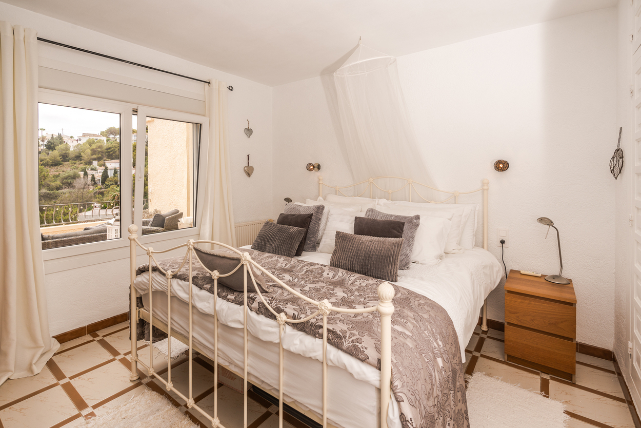 Slaapkamer met wit metalen bedframe, patroontegels en een raam met dubbele beglazing dat uitzicht biedt op de omgeving. Voorzien van een houten nachtkastje.